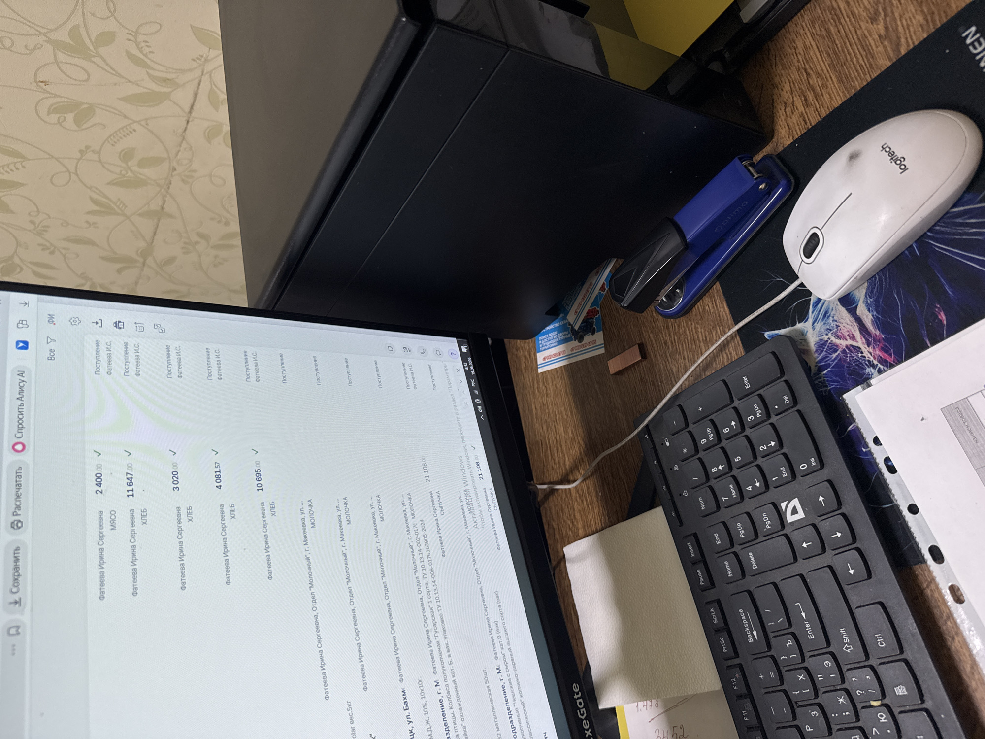 The image shows a close-up view of a computer monitor displaying a spreadsheet with financial data, a keyboard, a mouse, and a blue stapler on a wooden desk. The monitor shows lines of text with names, product types, and monetary values. Some lines also contain addresses and product descriptions. The date "19.06.2026" is visible at the bottom right of the screen. The background features a wall with a floral wallpaper pattern. The scene is likely an office or home workspace. There are no people visible in the image.
