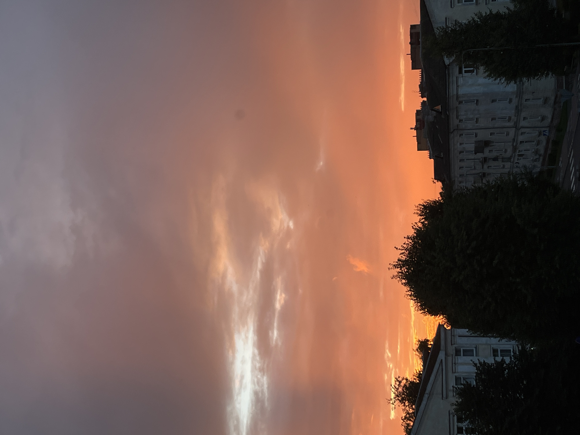 The image depicts a sunset over the city of Kohtla-Järve linn, Estonia. The sky is a dramatic display of orange, pink, and purple hues, with clouds catching the light. In the foreground, silhouetted against the vibrant sky, are the rooftops and upper floors of several apartment buildings. Trees, also dark silhouettes, frame the buildings and partially obscure them. The architecture suggests older, multi-story residential buildings common in urban areas. A street with a pedestrian crossing is visible in the lower right portion of the image. There are no people or distinct actions captured in the scene, which is primarily a landscape showcasing the natural phenomenon of a sunset over an urban environment.