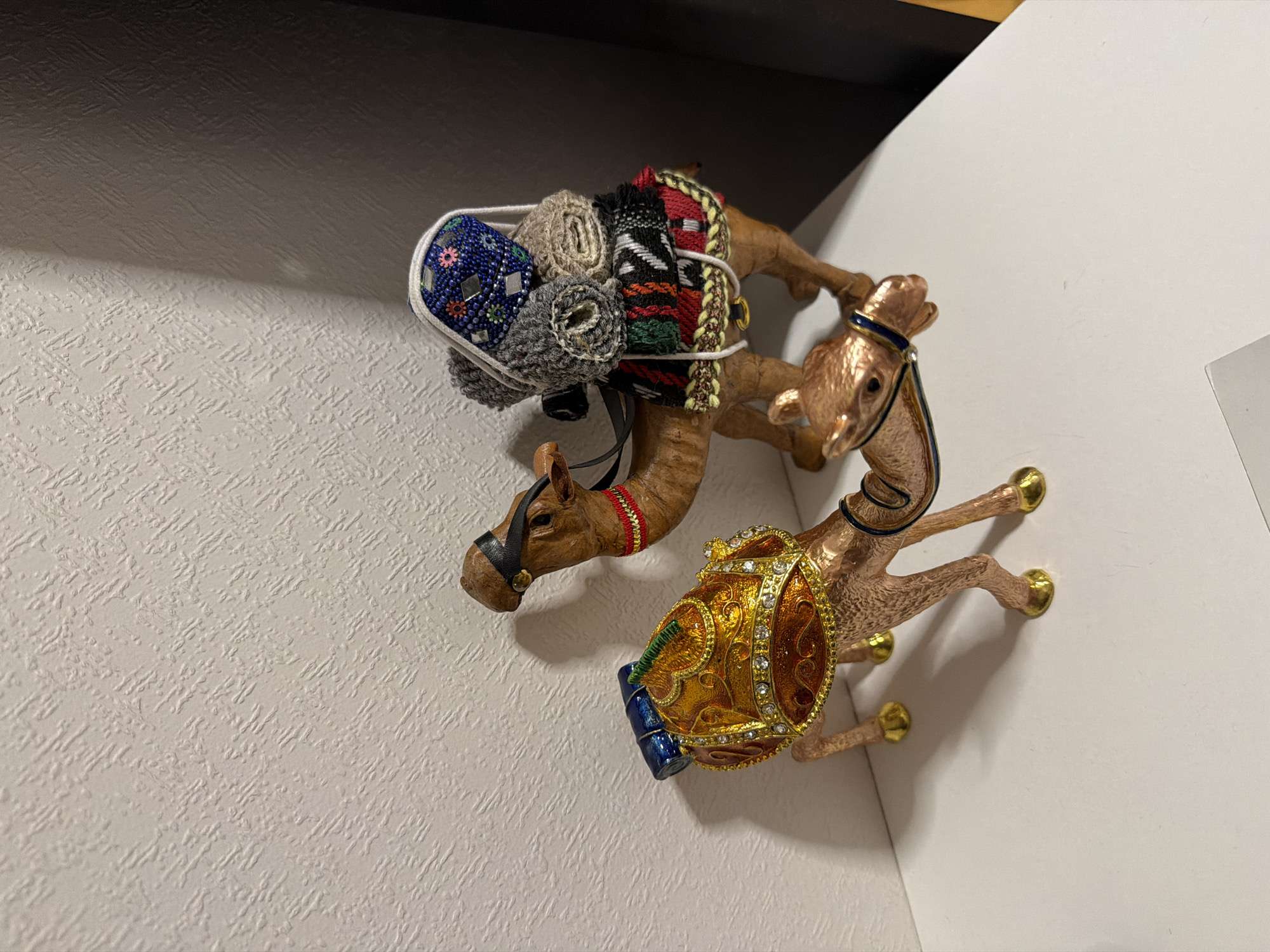 Two camel figurines are resting on a white textured surface. The camel on the left is brown and adorned with colorful fabric rolls and a beaded carrier on its back. The camel on the right is gold-toned and embellished with jewels and a blue accent. The background consists of a white wall and a portion of a dark-colored surface above. There is no indication of the city of Moscow, Russia.
