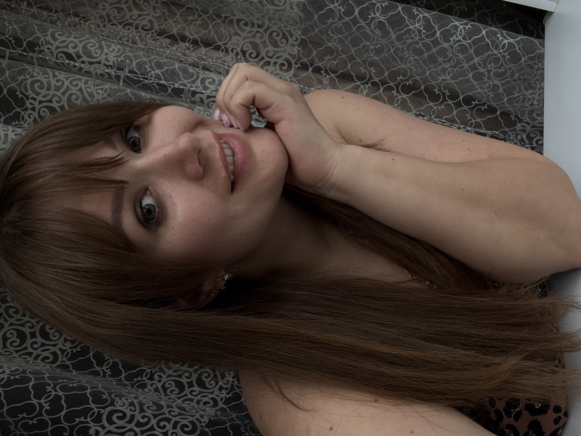 This is a vertical, medium shot photograph of a young woman with long, brown hair and bangs. She is lying down and looking at the camera with a slight smile, her hand resting on her chin. The background is a patterned curtain. The lighting appears to be indoor and soft. The woman has several piercings in her ear. The overall mood is relaxed and intimate. It is difficult to determine the time of day or specific location within Chaikovsky, Russia, from the image alone. There is no visible text in the photograph.