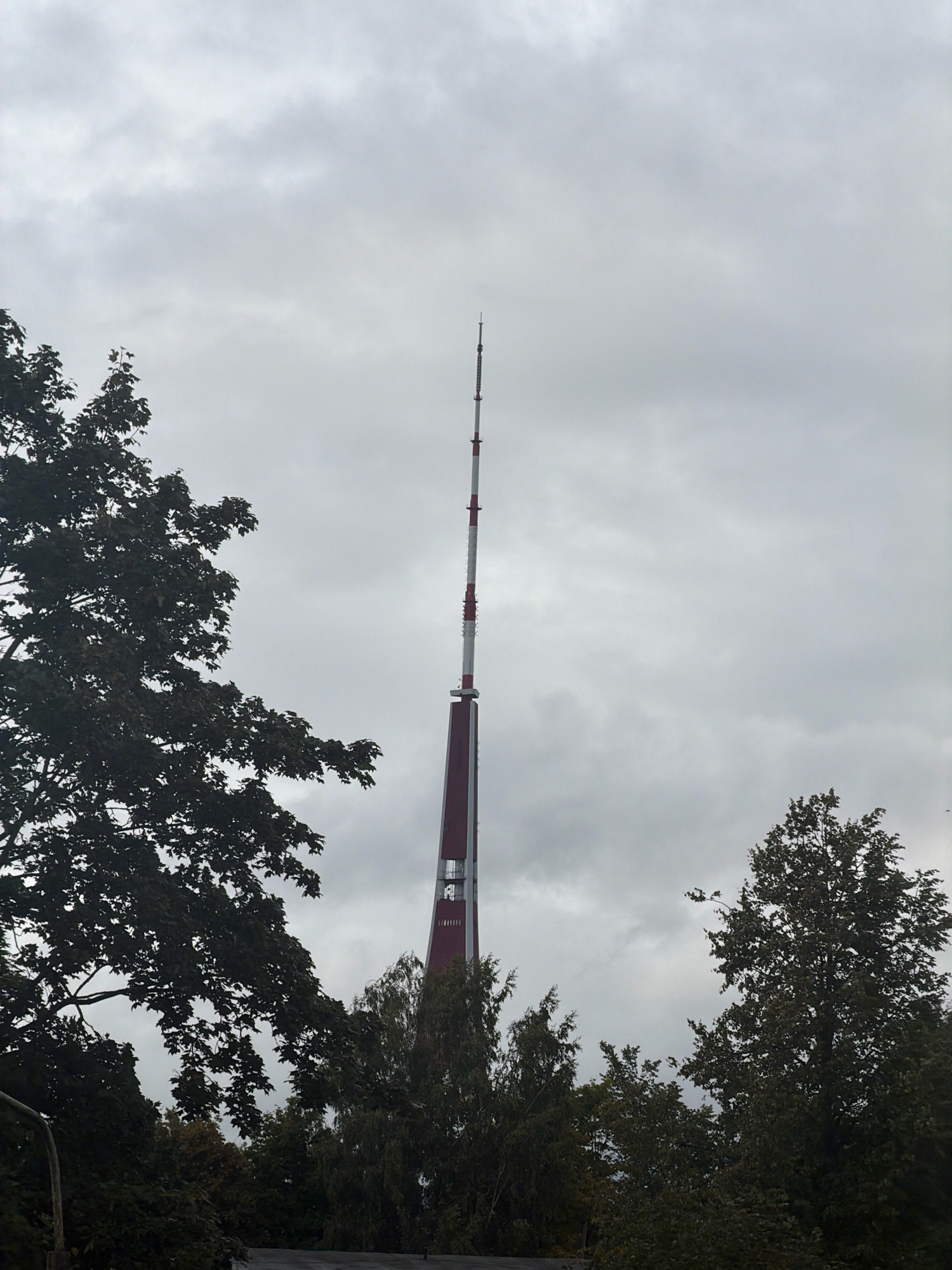 The image shows the Riga Radio and Television Tower, the tallest structure in Riga, Latvia, on a cloudy day. The tower, with its distinctive red and white markings, rises high into the overcast sky. In the foreground and to the sides, lush green trees obscure parts of the lower section of the tower and the surrounding landscape. The dense foliage suggests it is either late spring or summer. The overall atmosphere is one of a partly obscured, towering landmark amidst natural elements.