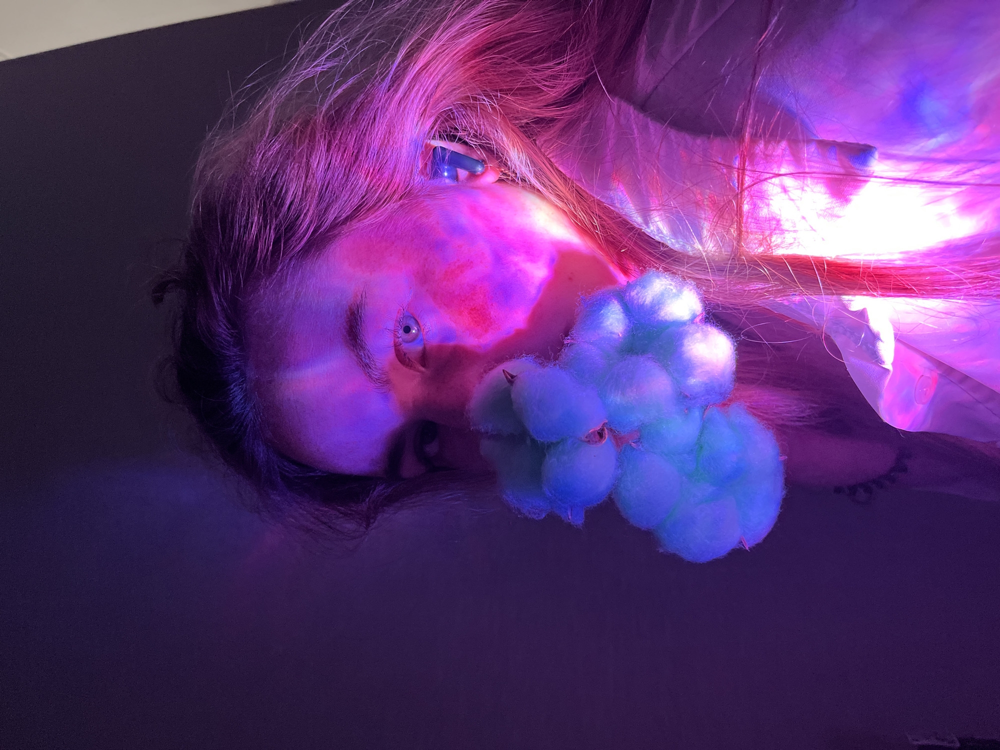 A woman lies on a dark surface, possibly the floor. She has fair skin and light-colored hair illuminated by pink and purple lighting. A cluster of blue and green cotton balls rests near her face. She is looking upwards, with one eye clearly visible. The exact location is unknown, and there is no evidence to suggest the location is Chaikovsky, Russia. The environment is dark and sparsely lit.