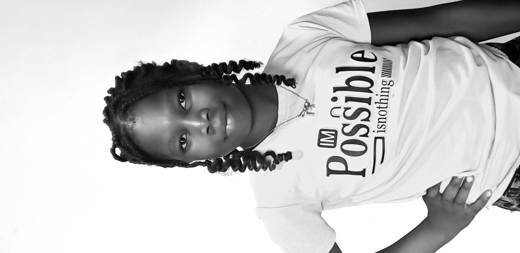 A young girl with braided hair and a bow necklace stands against a white background. She is wearing a white t-shirt with the words "IM Possible is nothing" printed on it. Her left hand is on her hip. The image is in black and white. The location is Sabon Gari, Nassarawa, Nigeria.