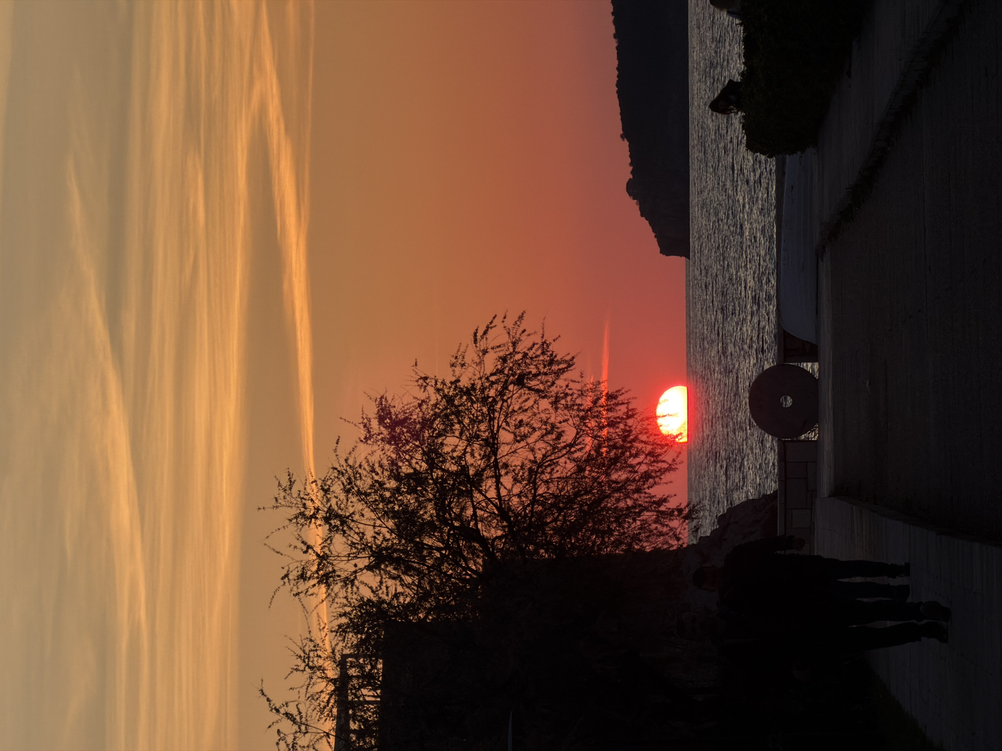 The image depicts a sunset scene over the sea in Sveti Stefan, Montenegro. The sky exhibits a gradient from orange-yellow with elongated, wispy horizontal clouds in the upper portion, transitioning to an intense red-orange closer to the horizon. A vibrant red-orange sun, partially submerged, rests on the water's surface, distinguished by a bright yellow-white band across its visible top edge. The sea presents a dark, choppy surface.
In the left foreground, a large, dark tree with discernible branches and sparse leaves is silhouetted against the sky, positioned adjacent to a dark, unidentifiable structure. Two silhouetted individuals walk away from the viewer on a paved pathway in the lower left. In the central midground, a dark, circular object resembling a millstone with a central aperture is situated next to a low, light-colored stone barrier that separates the pathway from the water. To the right, a dark, bushy hedge extends along the path, partially obscuring one silhouetted individual. The far right background features a dark, hilly landmass or island rising from the sea, topped by a small, indistinct structure. All foreground and midground elements are rendered as dark silhouettes due to the strong backlighting of the sunset.