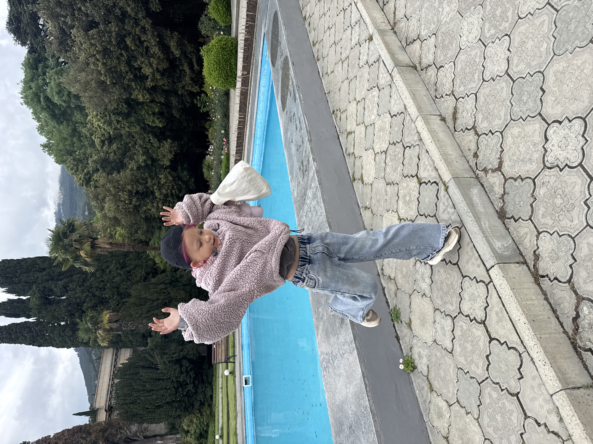 A child stands next to a rectangular, light-blue pool edged with grey concrete, against a backdrop of trees and partial mountain views. The child, wearing a pink, fluffy jacket, blue jeans, and a black beanie with a pink stripe, is positioned on grey, patterned pavers, arms outstretched. The sky is overcast. The city is Unknown, Unknown.