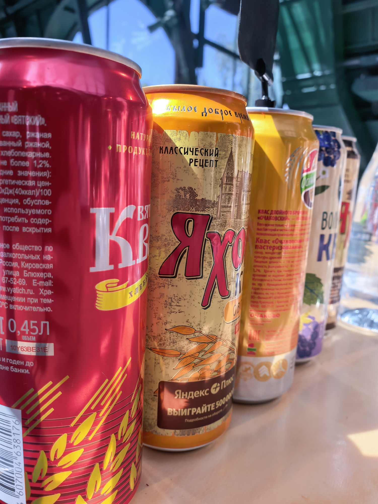 A close-up, eye-level, outdoor, medium shot captures a row of various beverage cans lined up on a light-colored surface. The lighting suggests a sunny day, casting shadows and highlighting the metallic sheen of the cans. In the background, blurred green structures and a bright blue sky are visible, hinting at an outdoor setting like a park or a market.
The foreground is dominated by a bright red can on the left, featuring yellow patterned designs and Russian text. Next to it, a golden can with an intricate design and Russian text is visible. To its right, a yellow can with Russian text and a circular emblem is present. Further down the line, parts of two more cans are visible, one with a purple floral pattern and another partially obscured.
The cans appear to be standard 0.45-liter aluminum beverage containers, likely containing non-alcoholic drinks given the visible text. No people are present in the image. The overall scene is clean and organized, with the focus squarely on the product display. The image was taken in Moscow, Russia.