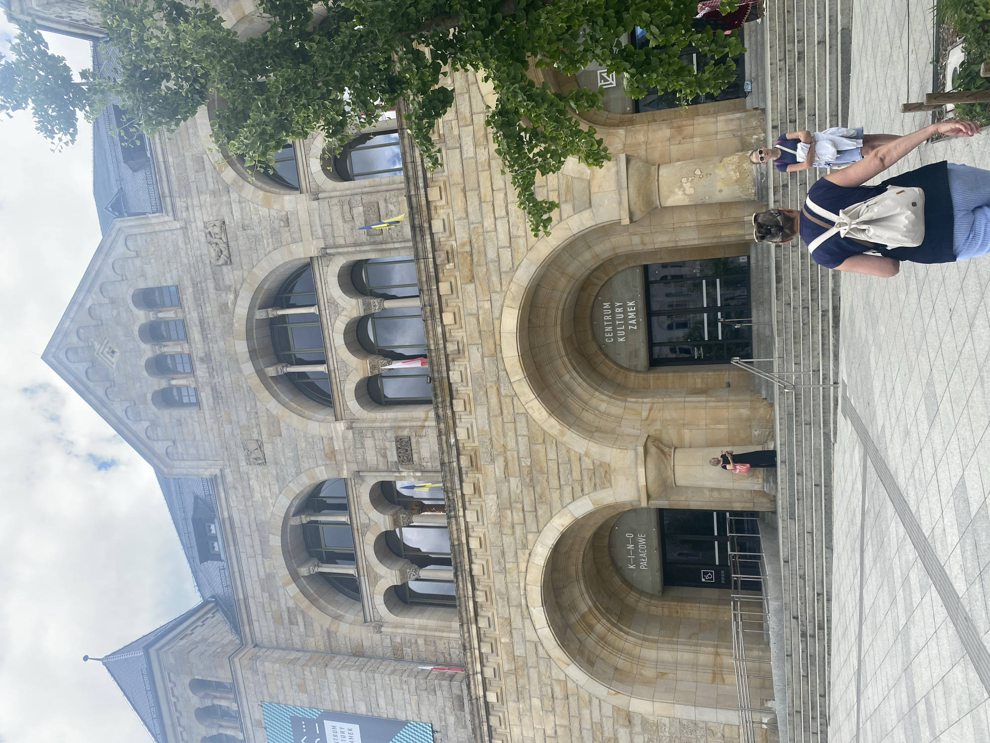 The image shows the exterior of the Imperial Castle (Zamek Cesarski) in Poznan, Poland. The grand building is made of stone, with large arched windows and entrances. Two individuals are walking up a wide set of stone stairs towards the building. One person in the foreground, wearing a dark blue t-shirt and striped pants, carries a cream-colored backpack. Another person, wearing a blue t-shirt and denim shorts, stands further up the stairs, holding a white jacket. A woman in a black dress stands to the left, near an entrance labeled "K-I-N-O PAŁACOWE." The building has flags flying from its facade, and a large green tree with fan-shaped leaves is visible to the right. The sky is overcast.