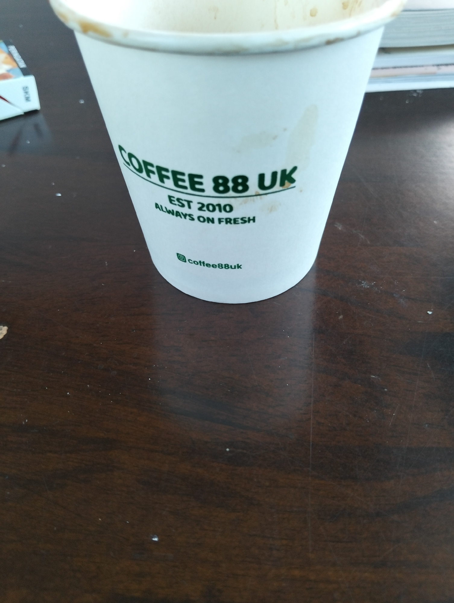 A close-up, eye-level shot shows a white paper coffee cup with green lettering that reads "COFFEE 88 UK EST 2010 ALWAYS ON FRESH". Below this, a small Instagram logo is visible, followed by "coffee88uk". The cup is positioned on a dark brown wooden table. To the left of the cup, a small portion of a white cigarette pack with red and blue accents is visible, with the word "SKM" printed vertically in black. The background is slightly out of focus, showing the edge of what appears to be a book or notebook. The lighting suggests an indoor setting, possibly a cafe or a home environment. The image composition is tight, focusing on the coffee cup and its immediate surroundings, with no discernible indications of the city of Julicotmeurak, Indonesia.