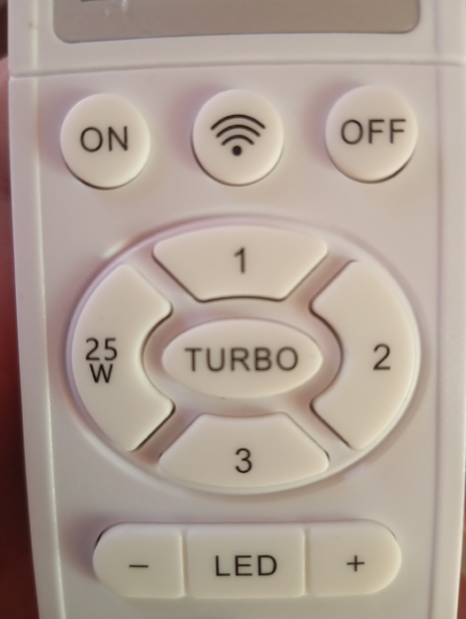 The image shows a close-up, eye-level shot of a white remote control. The remote has several buttons with text and symbols. At the top are three circular buttons labeled "ON", a Wi-Fi symbol, and "OFF". Below these is a circular arrangement of four buttons. One button is labeled "25 W", another "1", another "2", and another "3". In the center of this arrangement is an oval button labeled "TURBO". At the bottom of the remote are three rectangular buttons. The left button has a "-", the middle button is labeled "LED", and the right button has a "+". The remote is held by a person's hand, which is partially visible on the left side of the image. The background is blurred and appears to be indoors.