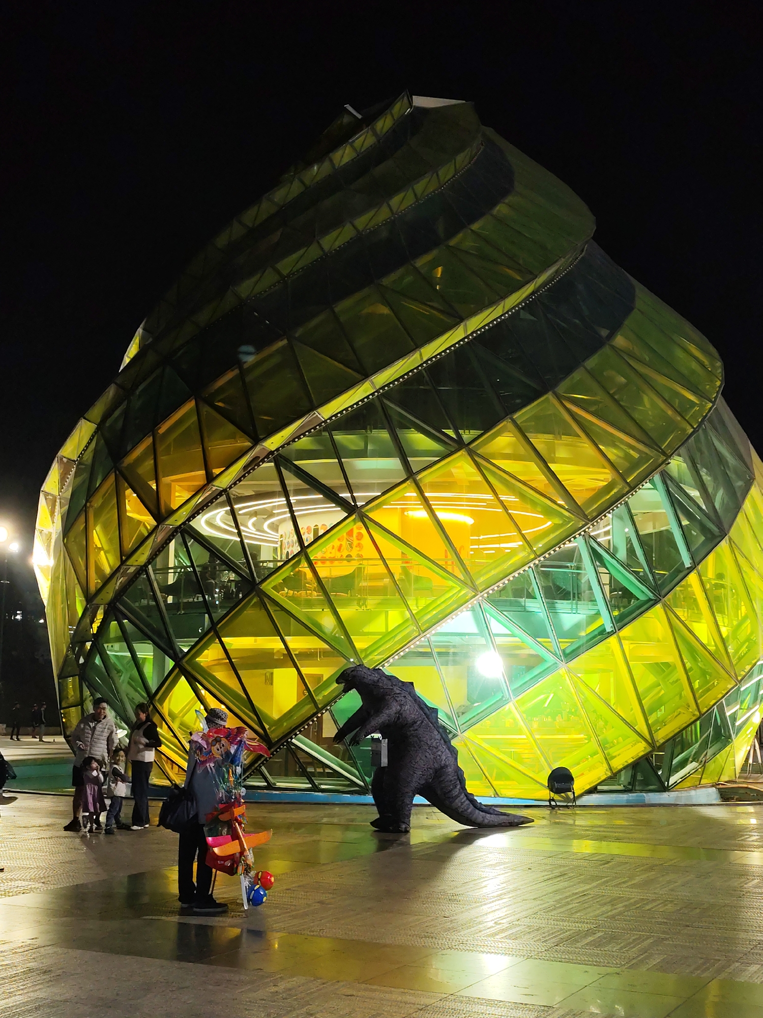 The image shows a nighttime scene in Da Lat, Vietnam, featuring the Da Lat Market. In front of the market is a life-sized Godzilla statue. To the left of the statue, a balloon vendor stands amidst a group of people, including adults and children. The ground is paved and wet. The market building itself is a large, uniquely-shaped structure with a geometric design and yellow and green lighting. The background is dark with minimal ambient light.