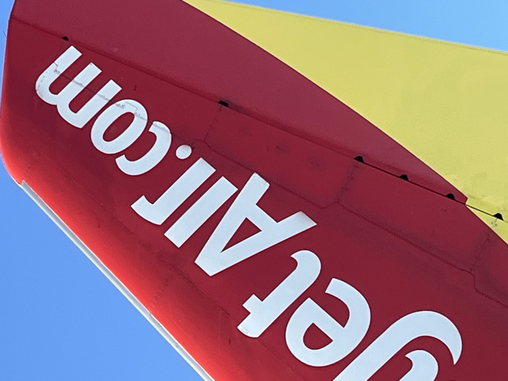 The image shows a close-up view of an airplane tail fin against a clear blue sky. The tail fin is painted red and features the white lettering "jetAir.com" written diagonally across it. A portion of a yellow stripe is visible along the bottom edge of the tail fin. The scene appears to be an airport or an area where aircraft are present. The specific location in Bac Cam Ranh Ward, Vietnam, is not discernible from the image itself, as the visual content focuses solely on the aircraft component. There are no people, actions, or interactions depicted.