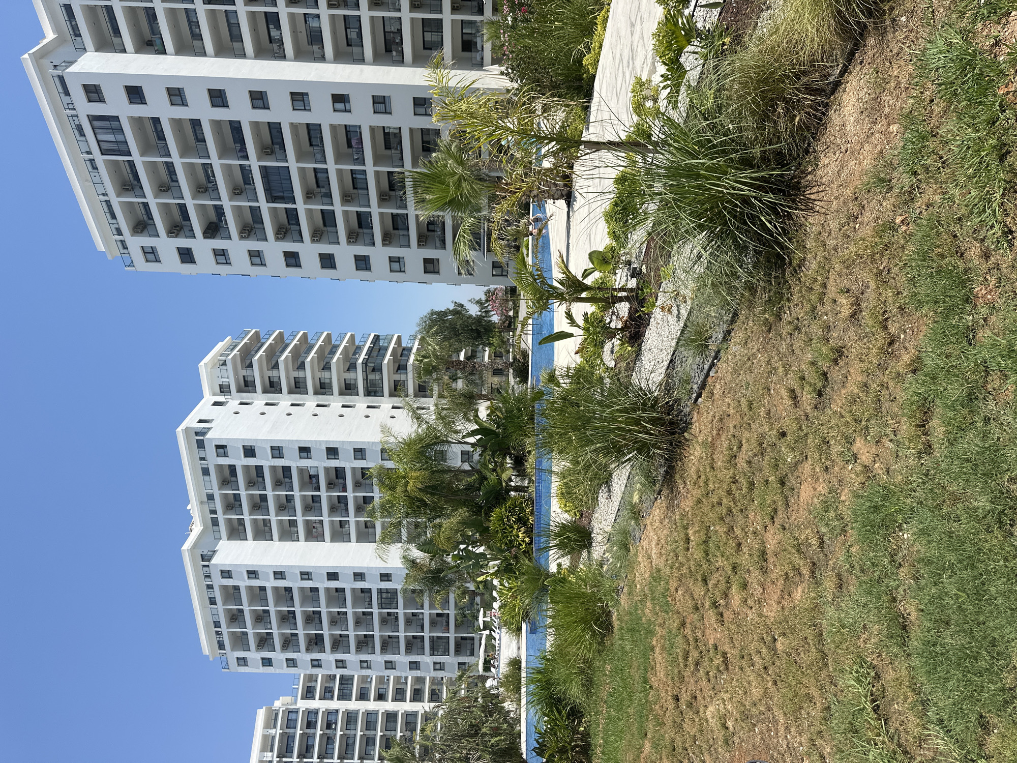 This media file is a vertical, eye-level shot of a modern resort complex in Trikomo, Northern Cyprus. The scene is dominated by several tall, white apartment buildings with repeating balconies and windows. In the foreground, a lush green lawn transitions to dry, brown grass, suggesting a sunny and potentially warm climate.
To the right of the buildings, a swimming pool with bright blue water is visible, surrounded by tropical plants and palm trees. Lounge chairs are scattered around the pool area, indicating a recreational setting. The sky is a clear, vibrant blue, suggesting a sunny day with no clouds.
The buildings appear to be residential or hotel accommodations, likely part of a resort designed for leisure and tourism. The architecture is modern and functional, with a focus on maximizing balcony space. The presence of air conditioning units visible on the balconies further emphasizes the warm climate.
The overall impression is one of a tranquil, sunny resort environment, likely during daytime hours. The landscaping, including the mix of healthy and dry grass, might suggest seasonal changes or a particular watering strategy for the grounds. No people are clearly visible in the image, and there is no visible text. The scene conveys a sense of relaxation and holiday atmosphere.