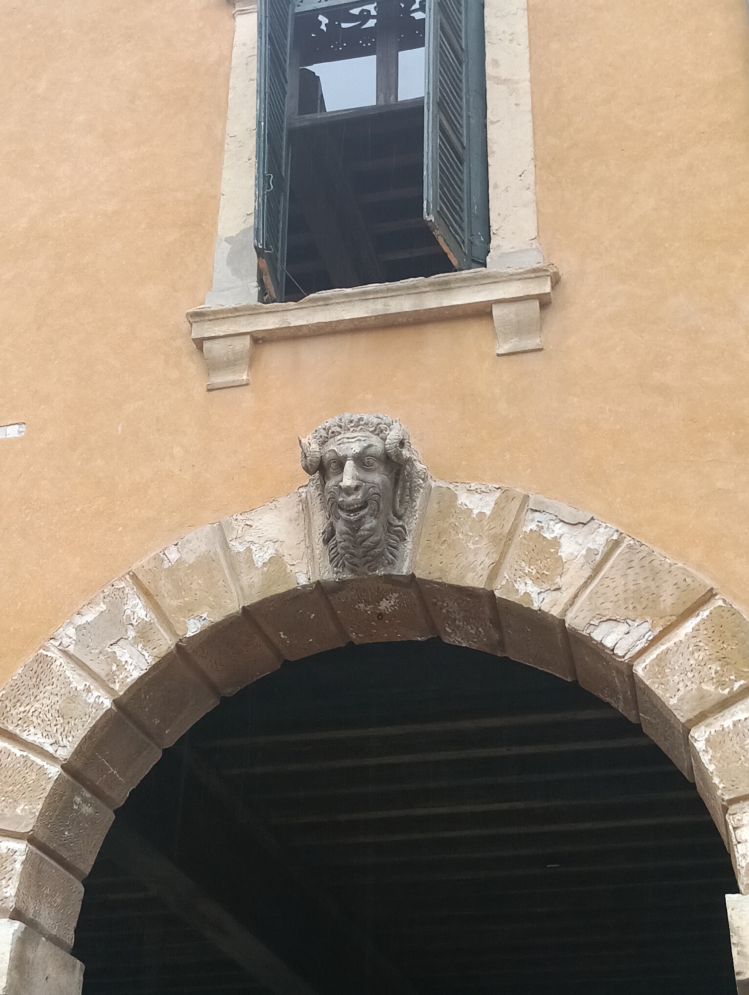 This is a detail of the facade of a building in Verona, Italy, likely located in the historic city center. The image focuses on a section of an ochre-colored wall featuring an arched stone entryway. Above the arch, a carved stone grotesque face with prominent features and curly hair is affixed. The arch itself is constructed from weathered, segmented stones, showing signs of age and wear. Directly above the grotesque, there is a simple, projecting stone ledge. Further up, a dark green shuttered window is visible, with one shutter ajar, revealing the darkness within. The overall impression is one of historical architecture, with elements of ornamentation and weathering typical of older structures in Verona.