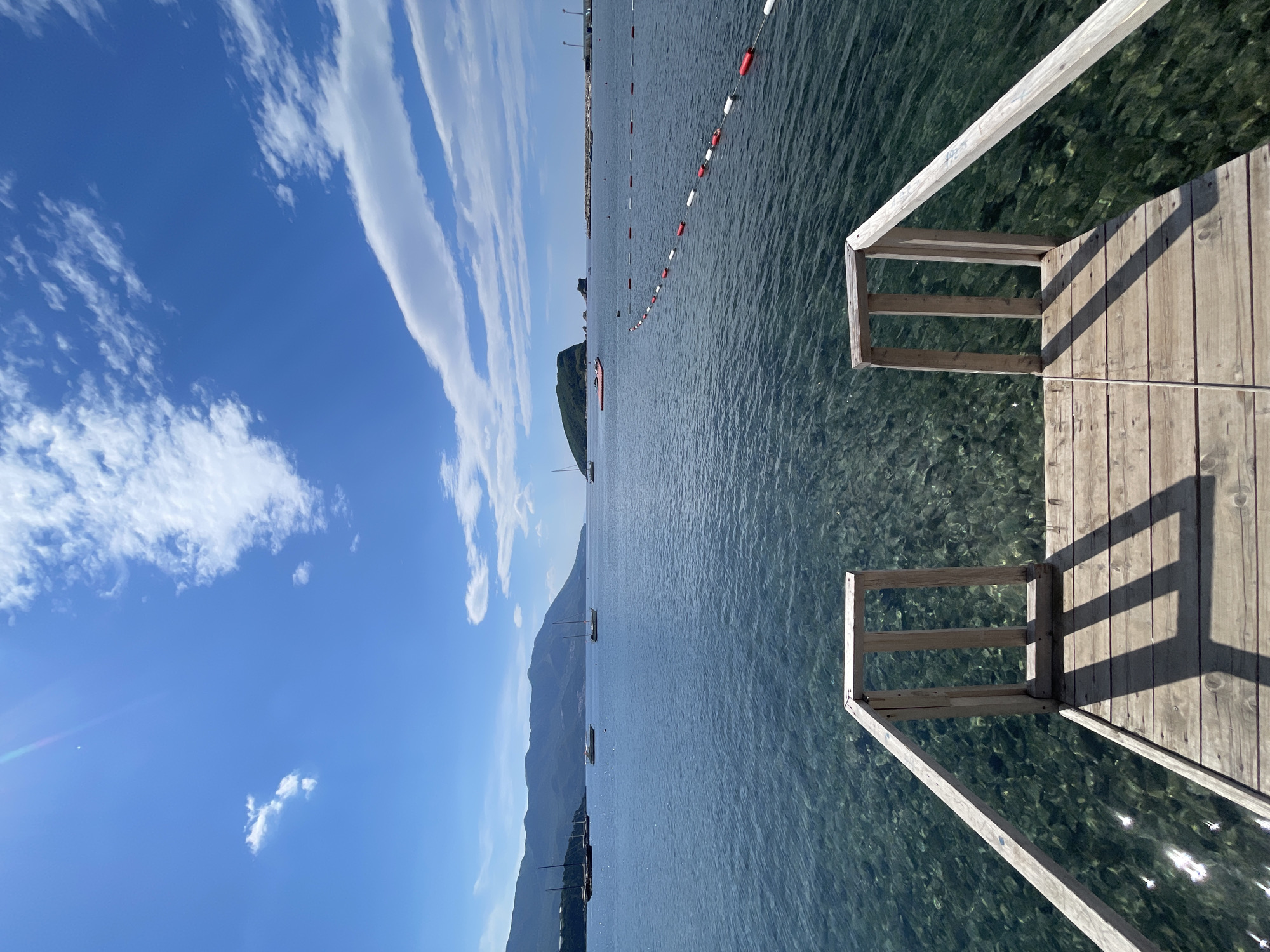 The image depicts a clear, sunny day at a waterfront location in Unknown, Unknown. A wooden pier extends into clear water, with a visible underwater view of the seabed. A rope line with red and white buoys stretches across the water, likely marking a swimming area. Mountains and islands are visible in the distance, with boats dotting the water's surface. The sky is blue with scattered clouds.