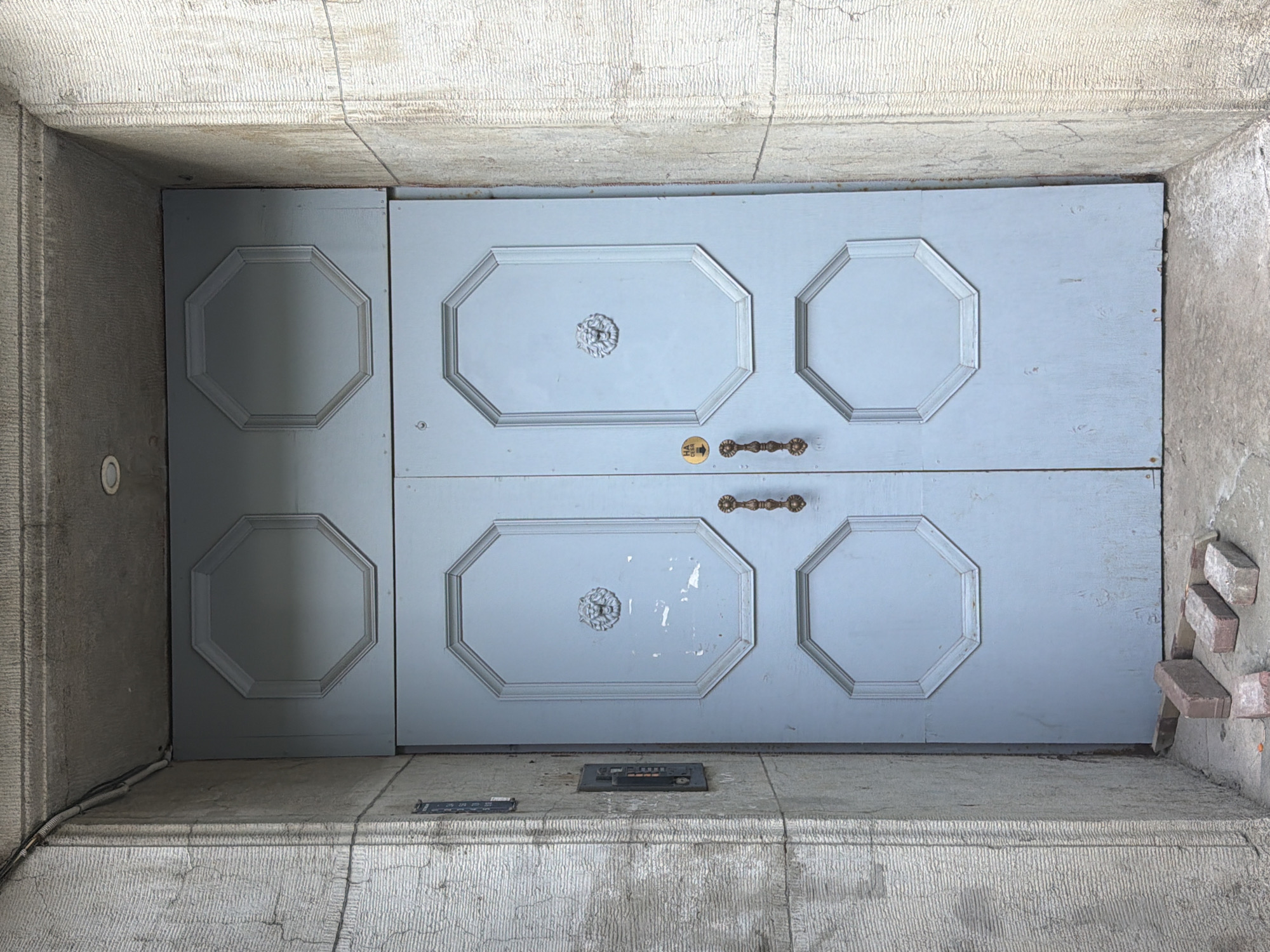 The image depicts a weathered, light blue double door, possibly an entrance to a building, located in Saint Petersburg, Russia. The doors are framed by rough, textured concrete walls. Each door has two octagonal panels, with the upper panels featuring decorative lion head medallions. The doors have ornate brass handles and a visible keyhole with a small yellow sticker to the left of it. On the left concrete wall, there are two dark intercom panels, one above the other, with visible buttons on the lower one. Three red and grey bricks are scattered on the ground in front of the doors, suggesting a state of disrepair or ongoing construction. The overall impression is of an old, somewhat neglected entrance in an urban setting.