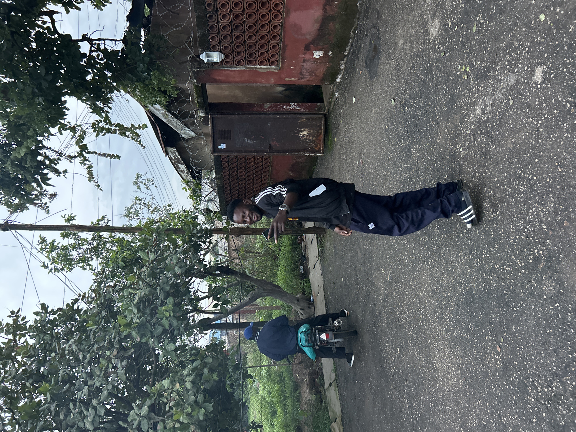 In Jos, Nigeria, a man in a black jacket with white stripes and navy blue track pants stands on an asphalt road, smiling and pointing. To his left, another man in a blue cap and jacket rides a small motorcycle. The background features a red building with an open metal door and decorative brickwork, barbed wire fencing, and trees with lush green leaves under an overcast sky.