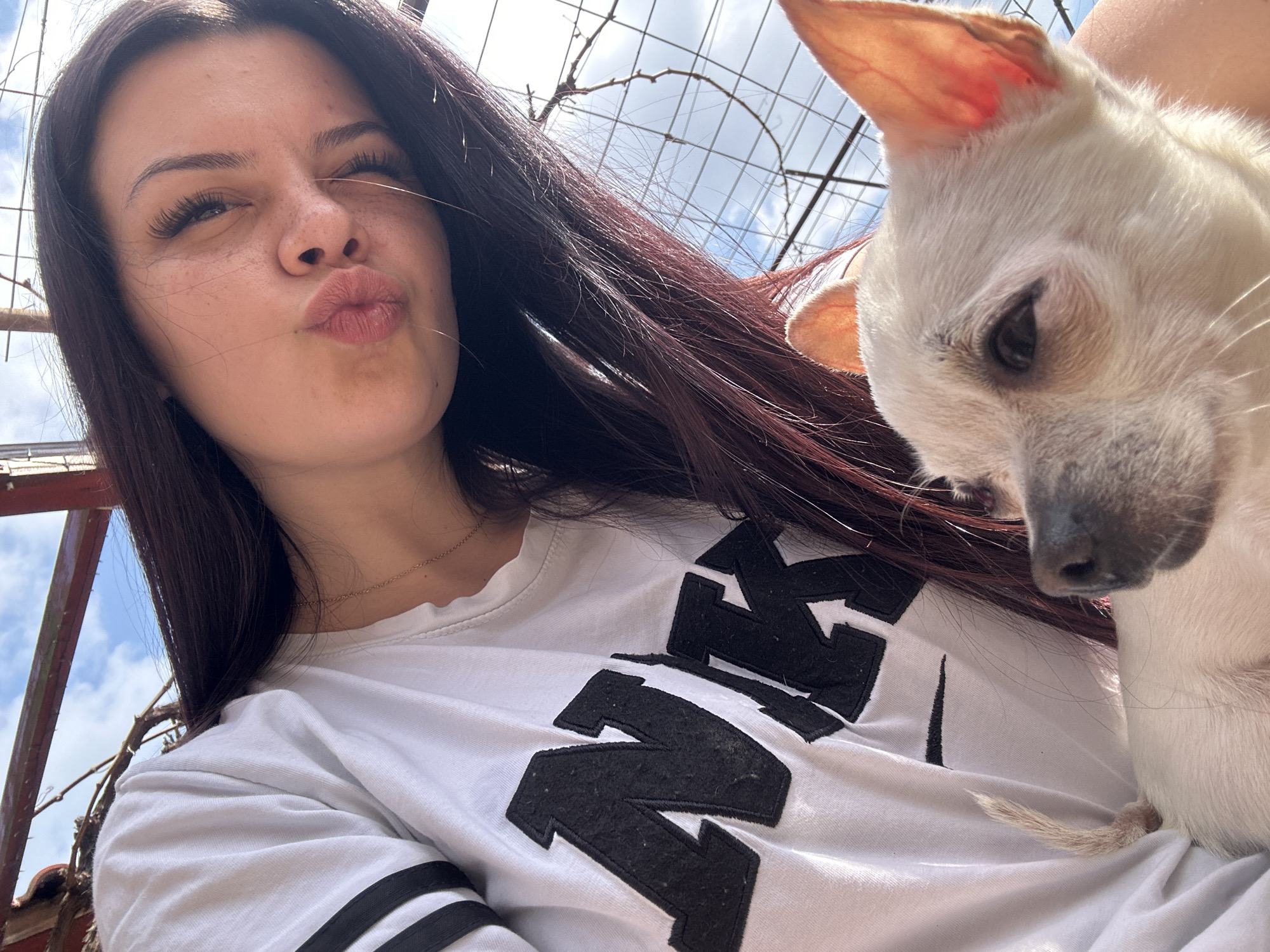 A woman with dark hair and freckles is posing for a selfie with a small, white dog. The woman is wearing a white t-shirt with a black logo on the front and is puckering her lips. The dog is nestled in her arms. Behind them is a metal fence, foliage, and a sky with some clouds. The location is Chubra, Bulgaria.