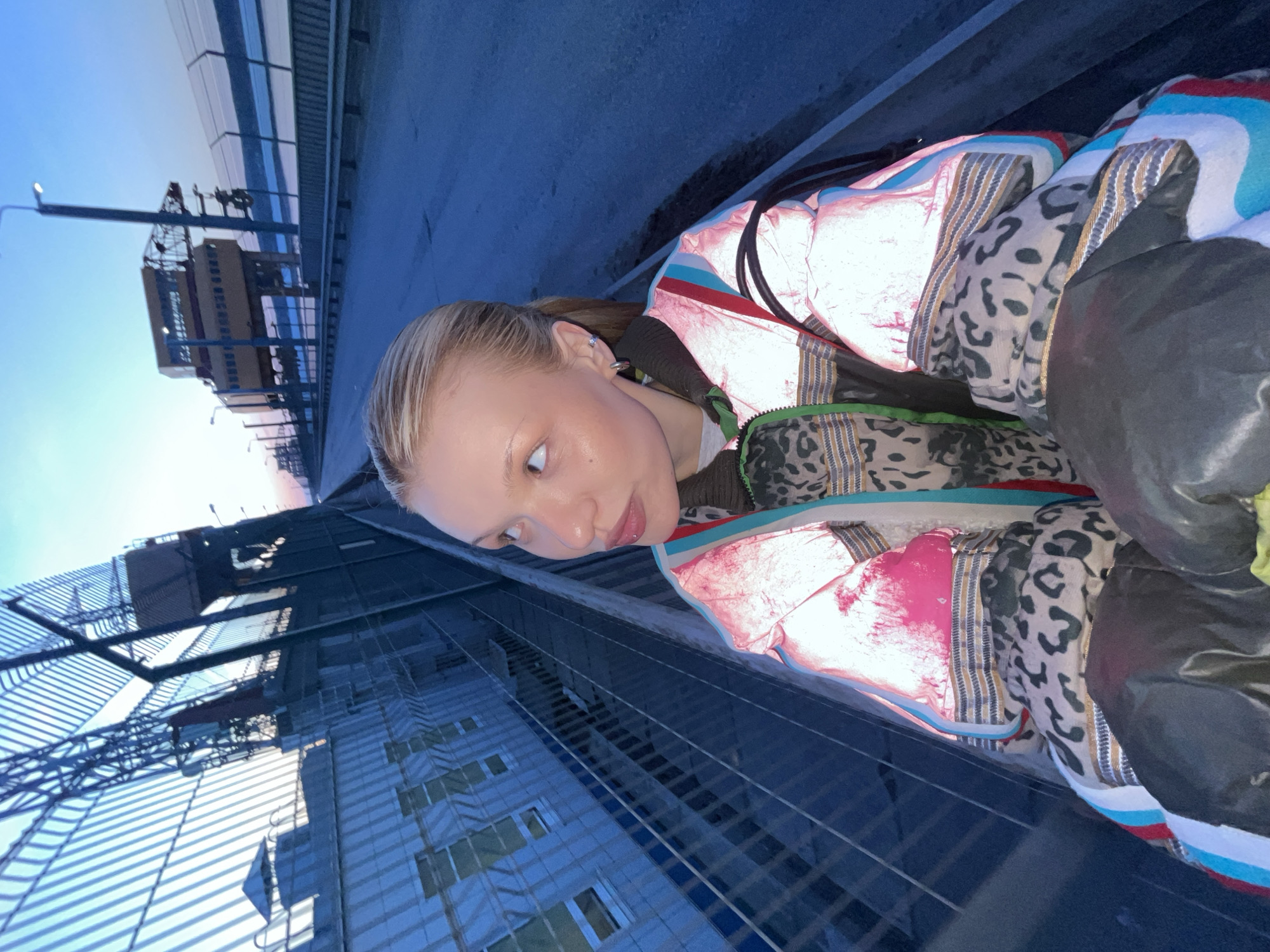 A full-body shot in a rotated low-angle perspective features a woman standing on an elevated structure, likely a bridge or rooftop, within the urban area of Chaikovsky, Russia. The woman has fair skin, blond hair pulled back, and wears a distinctive, multi-layered jacket featuring reflective material, animal print sections, and colored stripes. She is looking towards the left of the frame with a slightly pensive expression. The background showcases a large modern building with repeating patterns of windows and metal grating, suggesting an industrial or commercial complex. In the distance, a construction crane and additional buildings are visible against the backdrop of a twilight sky. The concrete structure of the elevated walkway or bridge is also part of the frame.