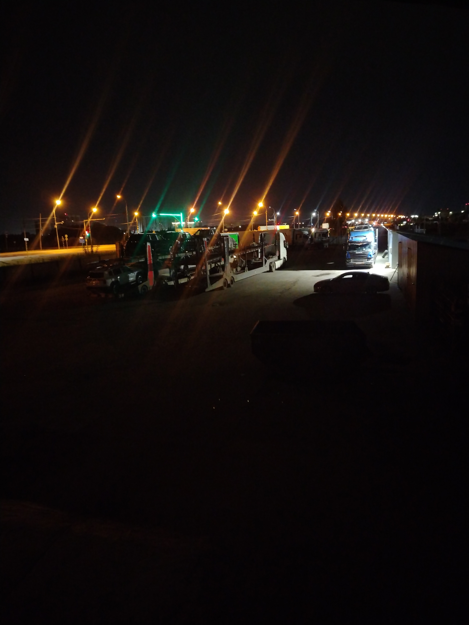 The image captures a nighttime scene at a transportation hub or parking lot in Krasnoglinnoye, Russia. Multiple large trucks, including a car carrier truck laden with vehicles, are parked. A blue semi-truck is also visible. A white sedan is parked in the foreground, illuminated by overhead lighting. The environment appears to be an industrial or commercial area, characterized by paved ground and the presence of buildings and streetlights. The lighting is artificial, cast by street lamps and vehicle headlights, creating long streaks of light across the dark sky due to a long exposure. The overall atmosphere is one of stillness and operation during night hours.