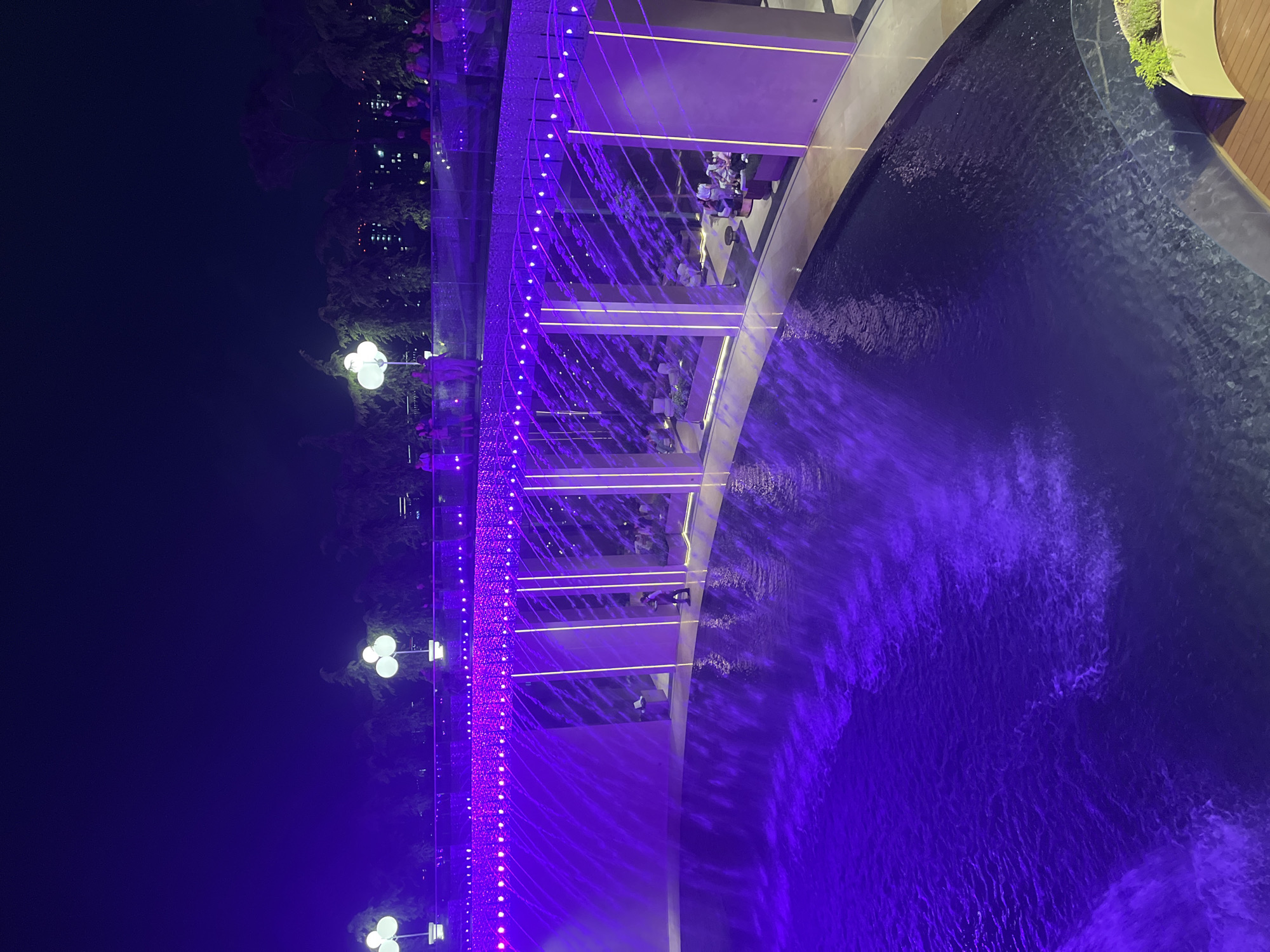 An elevated night-time, wide shot captures an outdoor lounge area with a water feature, illuminated by purple lights. A curved, elevated walkway lined with bright purple string lights and several globe-shaped lamps runs along the top of a structure. Below the walkway, a U-shaped water feature or pool is visible, with water cascading down the sides of the structure, creating a misty effect. The lower level of the structure features a lounge area with scattered seating arrangements, some occupied by people, and illuminated by vertical white light fixtures. A single person is visible on the lower level, walking near the edge of the water feature. The background shows a dark sky with trees and distant city lights, suggesting an urban setting. This location is likely in Krasnodar, Russia, specifically at a place like the "Solaris" complex, known for its illuminated water features and lounges.