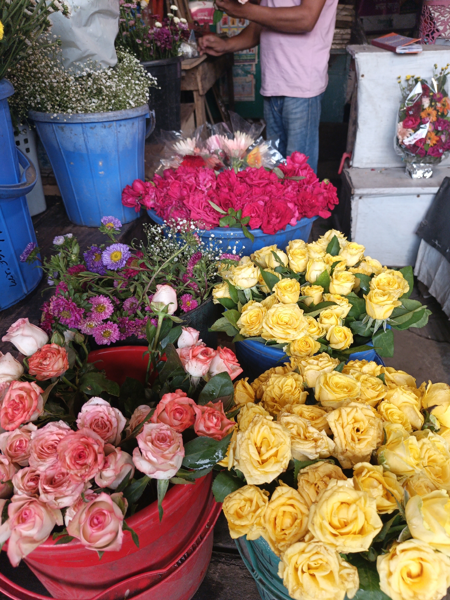 The image shows a vibrant flower market in Dhaka, Bangladesh. In the foreground, several colorful plastic buckets overflow with roses: pink, yellow, and red. Blue buckets hold baby's breath and other mixed flowers, including purple daisies. A man in a pink shirt and jeans stands behind the flower display, possibly arranging or selling flowers. A packaged bouquet is visible on a white surface in the background.