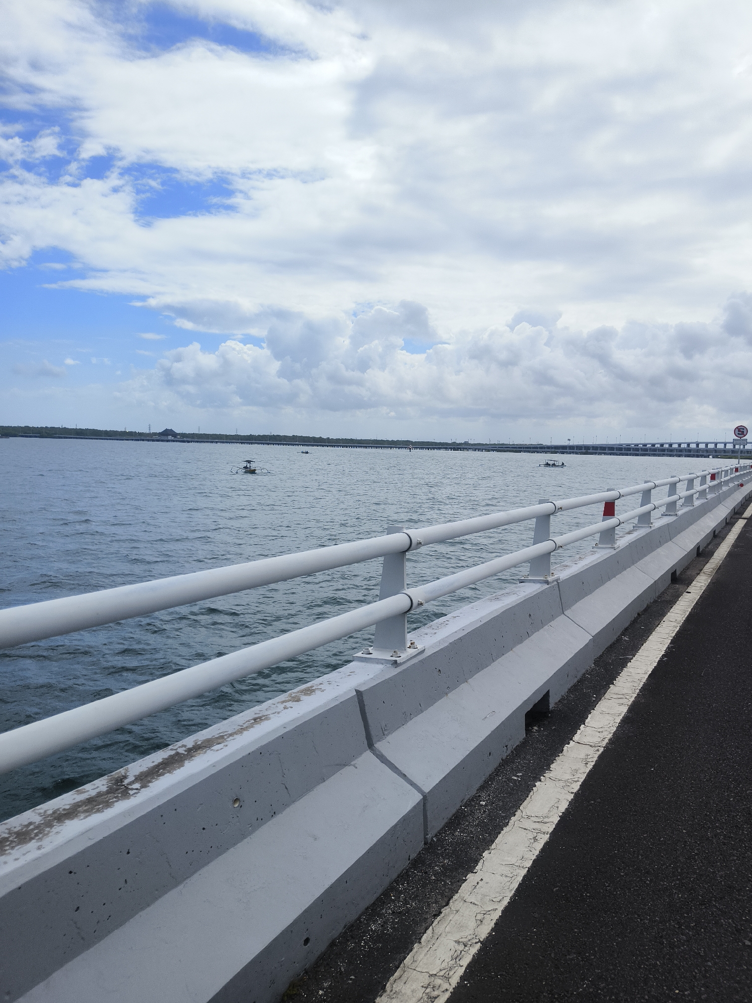 The image shows a view from a bridge overlooking a body of water, likely the Indian Ocean, near Kuta Selatan, Indonesia. The bridge features a white metal railing and a concrete barrier along its edge. The water is calm with some gentle ripples. In the distance, several small boats can be seen on the water. A long bridge structure extends across the water in the background, with what appears to be power lines or communication towers on the horizon. The sky is partly cloudy with a mix of blue sky and white clouds. The road surface of the bridge is visible in the foreground on the right, with a white line marking the edge. A speed limit sign, indicating 50, is visible on the right side of the bridge.