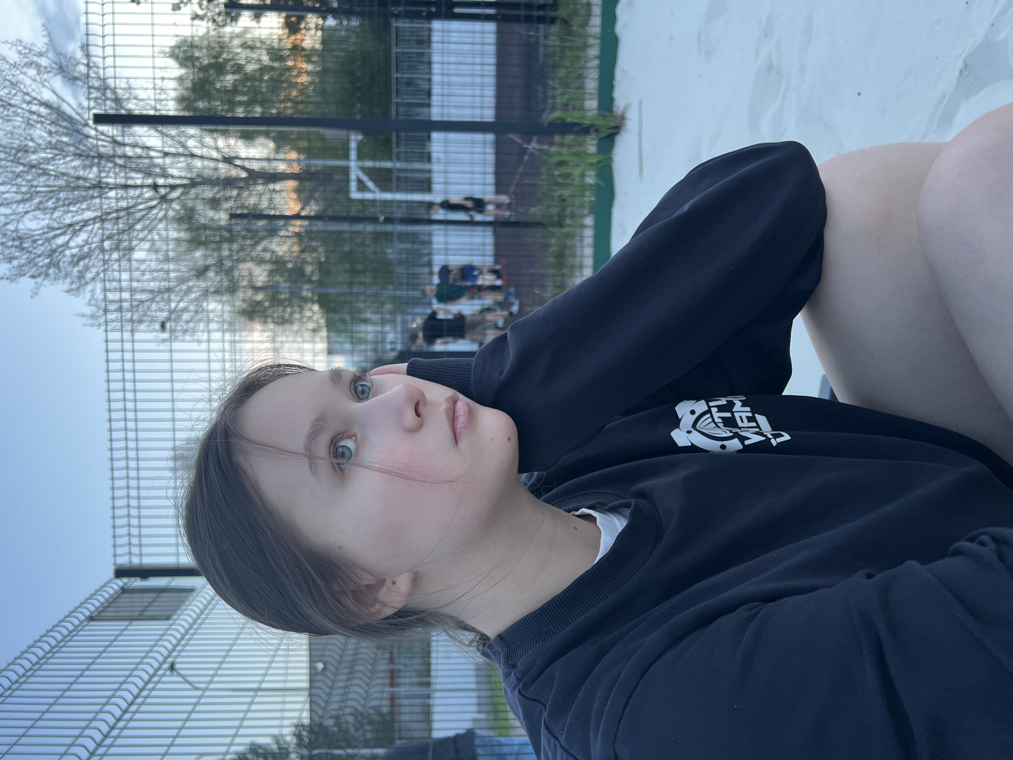 A young woman is the subject of this outdoor photograph. She has light skin, dark hair, and light-colored eyes. She is wearing a dark-colored sweatshirt. The woman appears to be sitting, with her arm raised and bent, positioned near her face.
In the background is a wire fence, a patch of green vegetation, and what appears to be a sports field or court with some people visible in the distance. The sky is visible above.
The location is identified as the city of Chaikovsky, Russia.