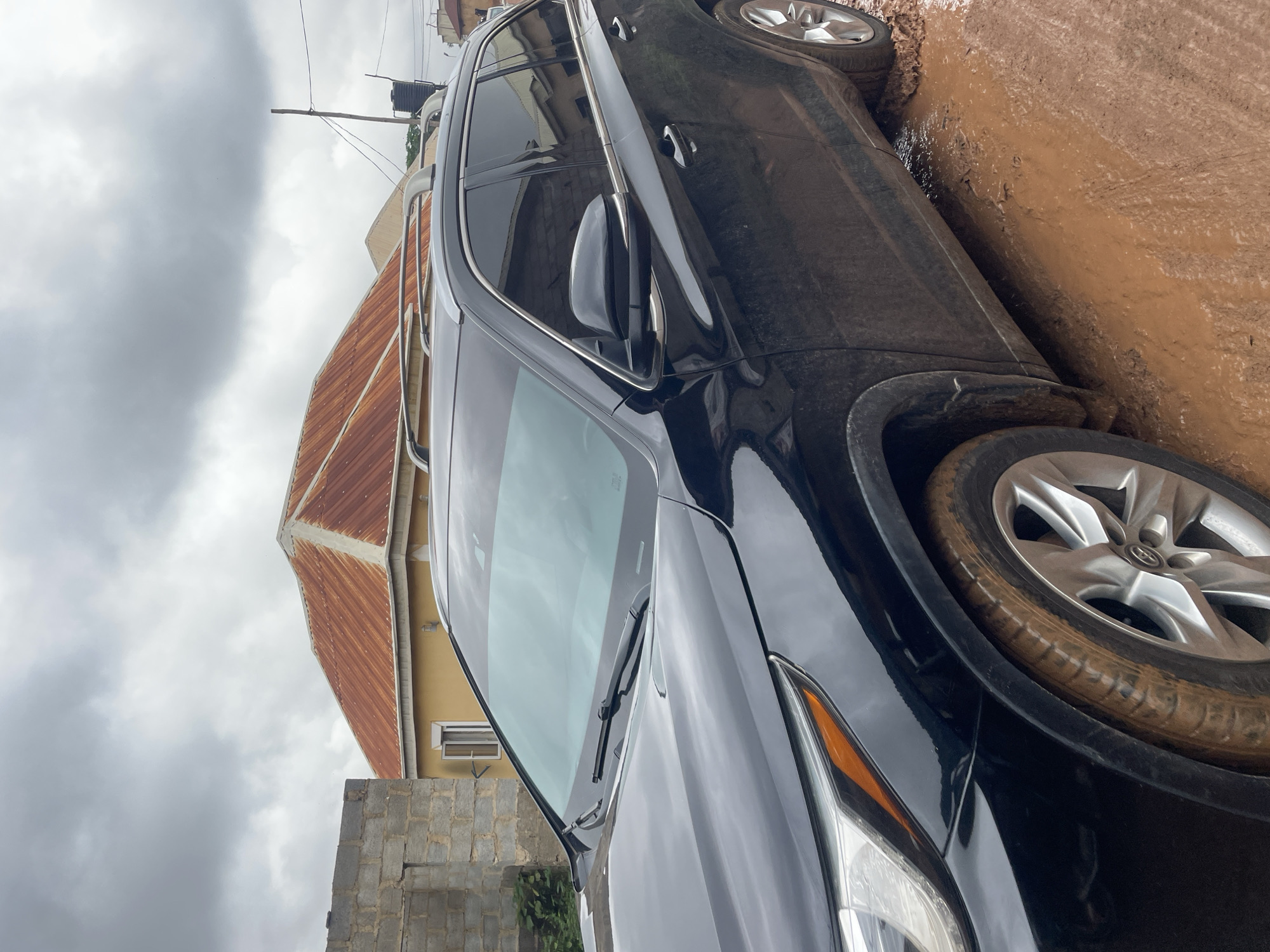 The image shows a black SUV stuck in thick mud on a dirt road in Kurunduma, Nigeria. The vehicle's front tires are partially submerged in the reddish-brown mud, with mud splattered on the car's body and wheel wells. The sky is overcast with heavy gray clouds. In the background, there are unfinished brick buildings and a house with a rusty corrugated metal roof. A water tank on a pole is also visible. The scene suggests a difficult driving condition, likely due to recent rainfall.