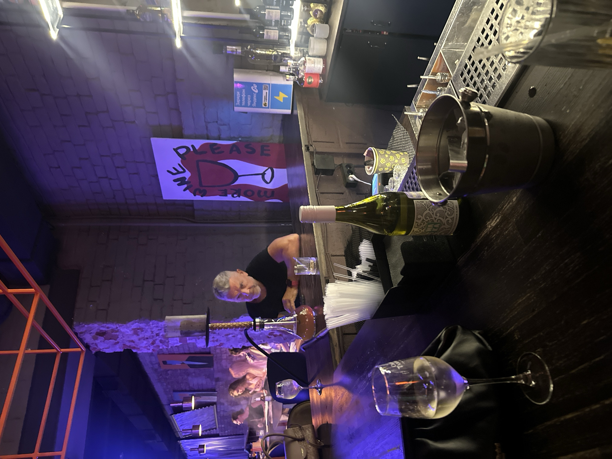 The image depicts an indoor scene, likely a bar or lounge, with a dimly lit atmosphere illuminated by purple and blue lights. In the foreground, a dark wooden bar counter is visible. On the counter, there is a wine glass filled with a light-colored liquid, a black purse, and a partially visible bottle of white wine. To the right, an ice bucket filled with ice and a drink in a glass with a straw are present.
Further back, a man with gray hair and a beard is seated at the bar, wearing a black t-shirt. He is looking down and appears to be interacting with something on the counter. A hookah is positioned behind him, with its base filled with amber liquid. In the background, several other patrons are seated at tables, some of whom appear to be looking at their phones. The walls of the establishment are made of brick, with some exposed rough plaster sections. A framed picture with a stylized image of a wine glass and text that reads "WINE PLEASE" is hanging on the wall. Shelves with various bottles of alcohol are visible behind the bar. The overall ambiance suggests a relaxed evening setting. There are no visible indications that this scene is located in Barnaul, Russia.