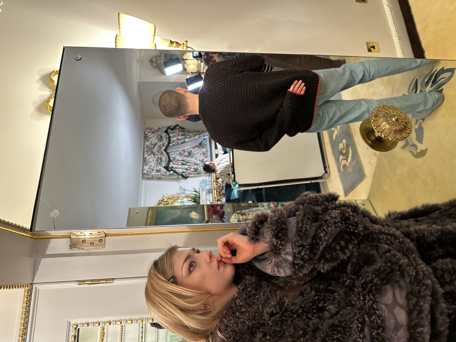 A mirror reflects a complex scene. A woman in a dark fur coat and blond hair looks left. The reflection shows a man in a dark sweater, light jeans, and a shaved head. Further back in the reflection, another man is visible. Lighting equipment is present, suggesting a photoshoot. The room features ornate decor, including floral curtains and gilded furniture. A brass doorknob and patterned carpet are visible on the floor.