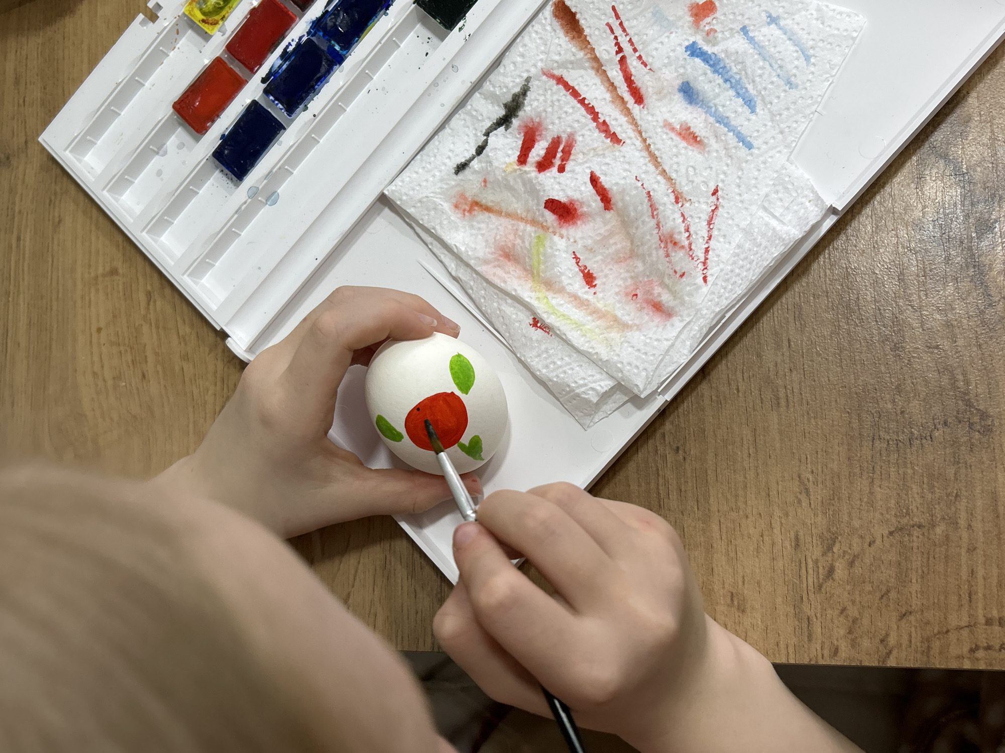 The image shows a person painting an egg. The person is holding the egg with their left hand and painting it with a brush in their right hand. The egg is white with a red circle and green leaves painted on it. There is a watercolor palette with different colors of paint to the left of the egg and a white paper towel with dabs of paint on the right. The surface below is made of wood. The image does not provide any information about the location being Chaikovsky, Russia.