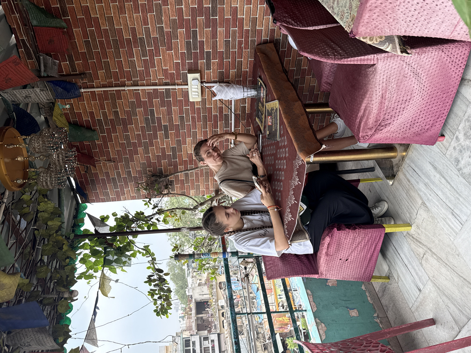 Two young women are seated at a table on an outdoor balcony overlooking a bustling street scene in Delhi, India. The woman on the left, with braided hair, is focused on her phone. The woman on the right, with her hand on her forehead, is looking towards the camera. The balcony is decorated with prayer flags and leafy plants, and the brick wall behind them suggests a rooftop cafe or restaurant. The street below appears to be a vibrant marketplace, with colorful awnings and a general sense of activity. The overall atmosphere is one of relaxed enjoyment amidst the energetic urban environment of Delhi.