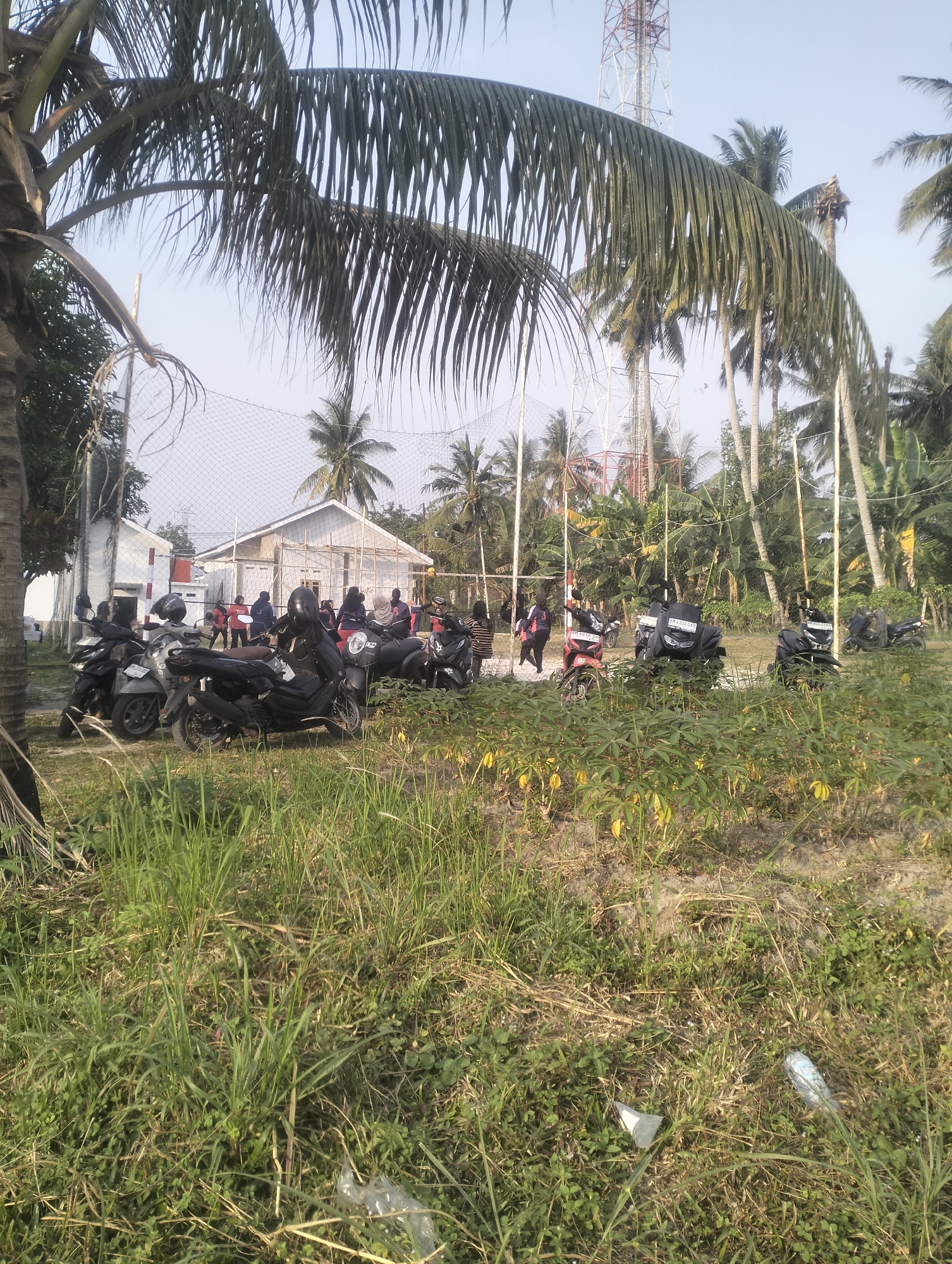 The image depicts an outdoor scene in Air Jamban, Indonesia, featuring a collection of motorcycles parked in front of a building. Several people are visible, some standing near the motorcycles and others gathered further back. The environment is characterized by abundant vegetation, including palm trees and tall grass. A volleyball net is set up in the background, and a telecommunications tower is visible in the distance. The sky is clear and bright. The overall impression is that of a communal gathering or event taking place in a tropical setting.