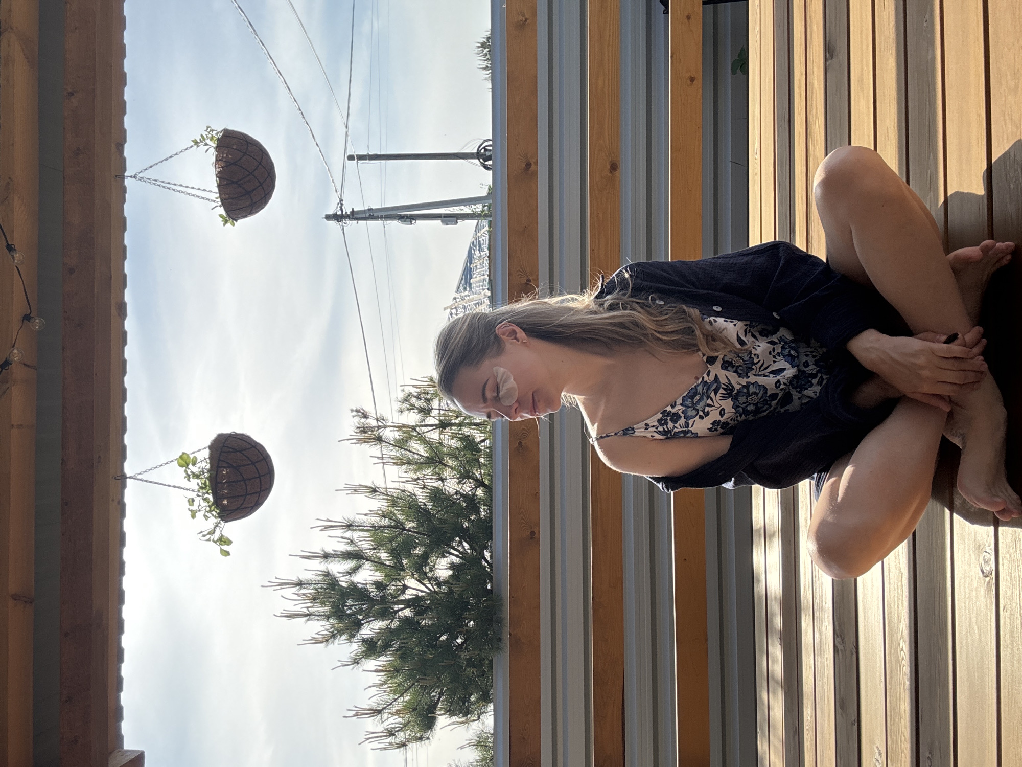 The image shows a woman sitting on a wooden deck. She is wearing a floral dress and a dark jacket. She is fair-skinned with blonde hair. She is sitting with her legs crossed and hands clasped. Behind her is a fence made of alternating wood and metal slats. Hanging above are two spherical planters. In the background are trees and power lines. The location is Chaikovsky, Russia.