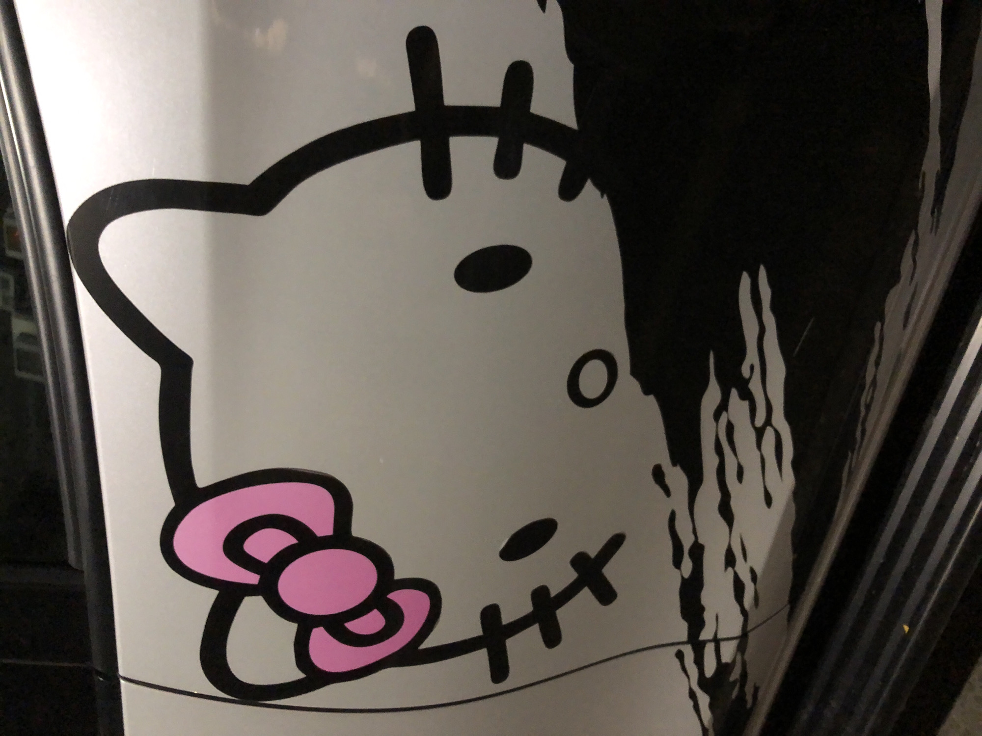 This media file shows a close-up view of a stylized cartoon character, resembling Hello Kitty, on a white surface. The character is depicted in black and pink. The left side of the image features the character's head, with its outline, two black oval eyes, and a pink bow on its left cheek. The character's mouth is represented by a horizontal line with three vertical strokes.
To the right of the character's head, there's a black, drippy, abstract design that partially obscures the character. This design appears to be a liquid or paint-like element, adding a contrasting texture and visual effect. The white surface the character is on seems to be curved, suggesting it might be part of an object like a vehicle or appliance. The background is dark, with some vertical striped elements visible on the far right, possibly indicating a structural element or another object.
There is no discernible text in the image. The lighting is somewhat dim, and there are no clear indicators of the time of day or weather conditions. The overall impression is of a decorative sticker or graphic design applied to a surface. The location context provided is Đà Nẵng, Vietnam, but this cannot be visually confirmed from the image itself.