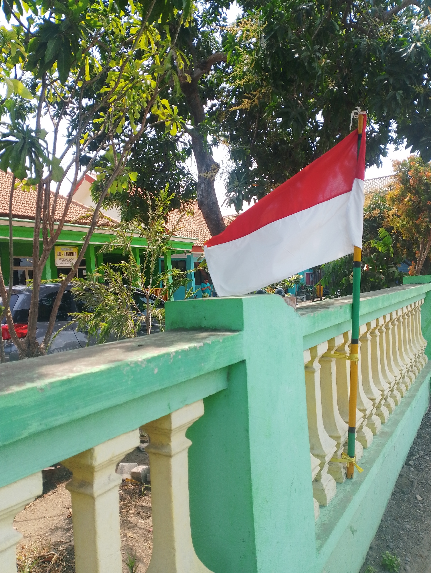 The image depicts an Indonesian flag, red and white, flying on a flagpole attached to a light green and cream-colored fence. The fence has decorative balusters and overlooks a dirt and gravel path. Behind the fence, a low-rise building with a terracotta tile roof and light green walls is visible. Trees with green foliage are scattered throughout the scene, casting shadows. The building has a sign that reads "MUSHOLLA AB-RAKHMAD" and other Indonesian text. A dark-colored car is parked in front of the building. The sky is bright, suggesting daylight. The location is identified as Suruh, Indonesia.