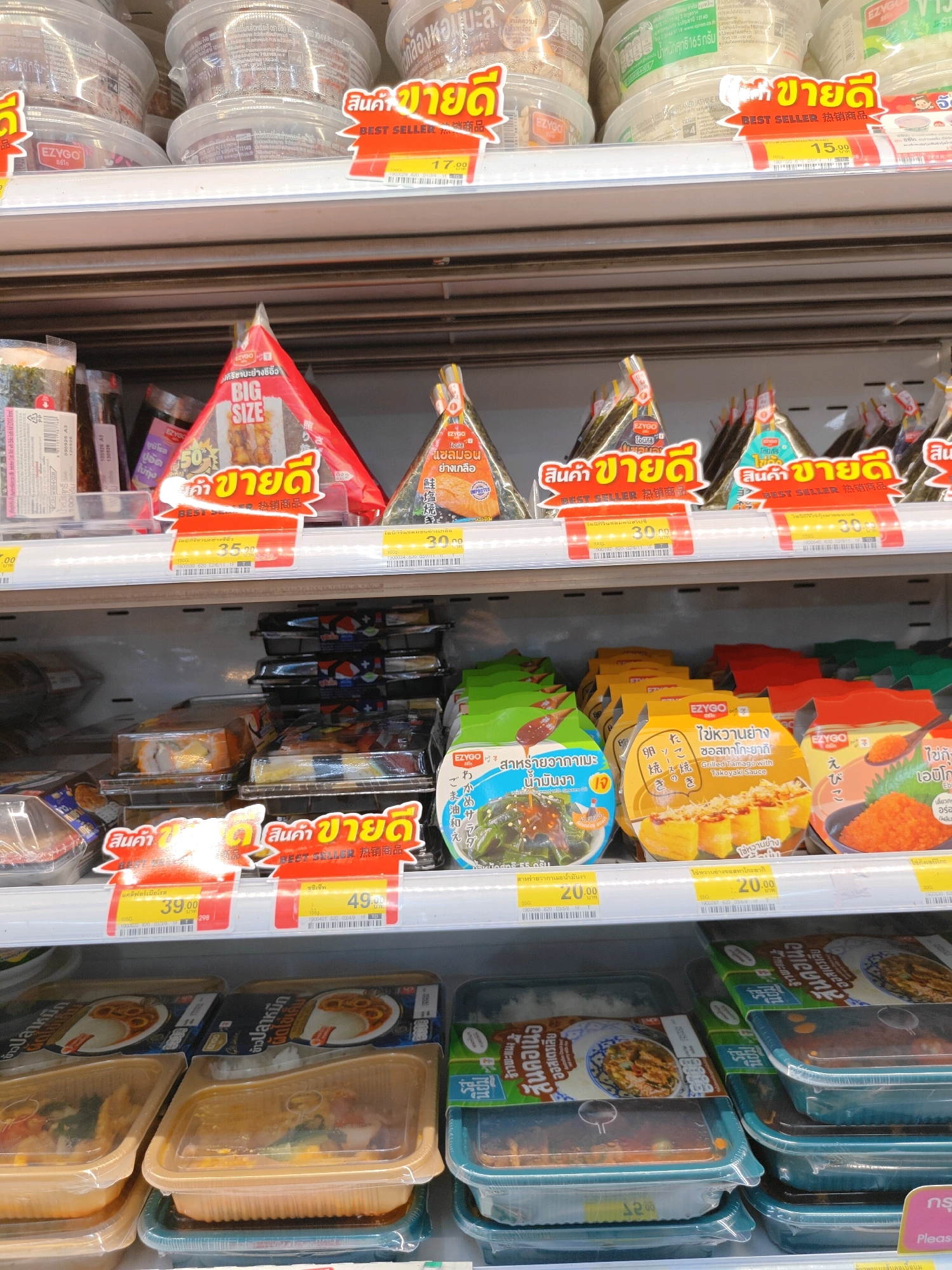 The image shows a retail shelf displaying various food products, likely in a convenience store or supermarket in Mu Si, Thailand. The products are packaged in plastic containers and bags, with prominent signage indicating they are "Best Seller." One section of the shelf features triangular rice balls, some labeled "Big Size." Another section displays pre-packaged meals, including what appears to be sushi and rice dishes. The overall environment is brightly lit, characteristic of a retail setting. No people are visible in the image.