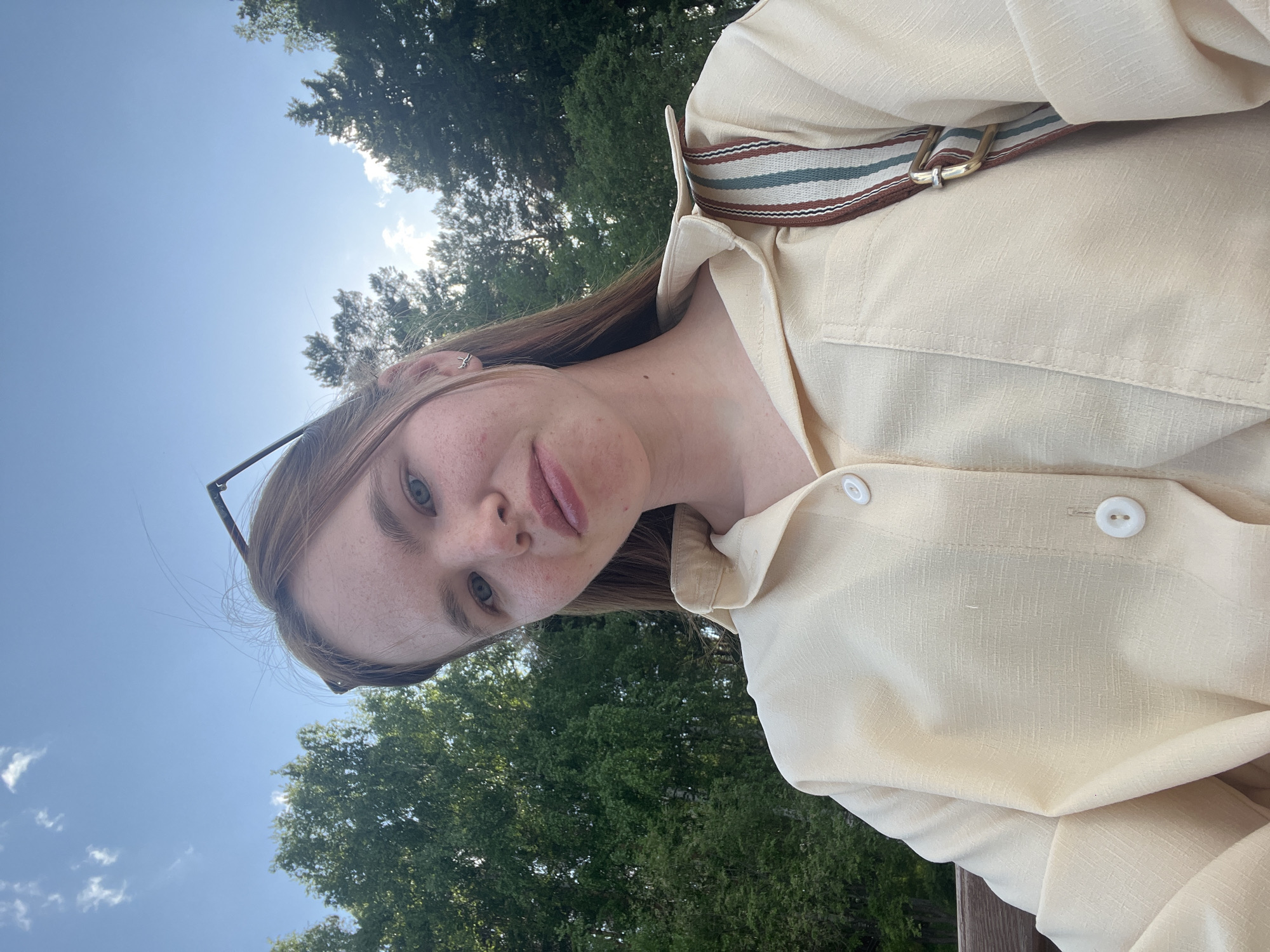 A light-skinned individual with freckles, brown hair, and blue eyes is centered in the frame, wearing a light yellow collared shirt and a strap with brown, blue, and white stripes running across the upper chest. The subject is outside during the day, with a blurred background of green trees and a blue sky with light clouds. The image is taken from a low angle, looking upward. The specific location within Chaikovsky, Russia, cannot be determined from the image.