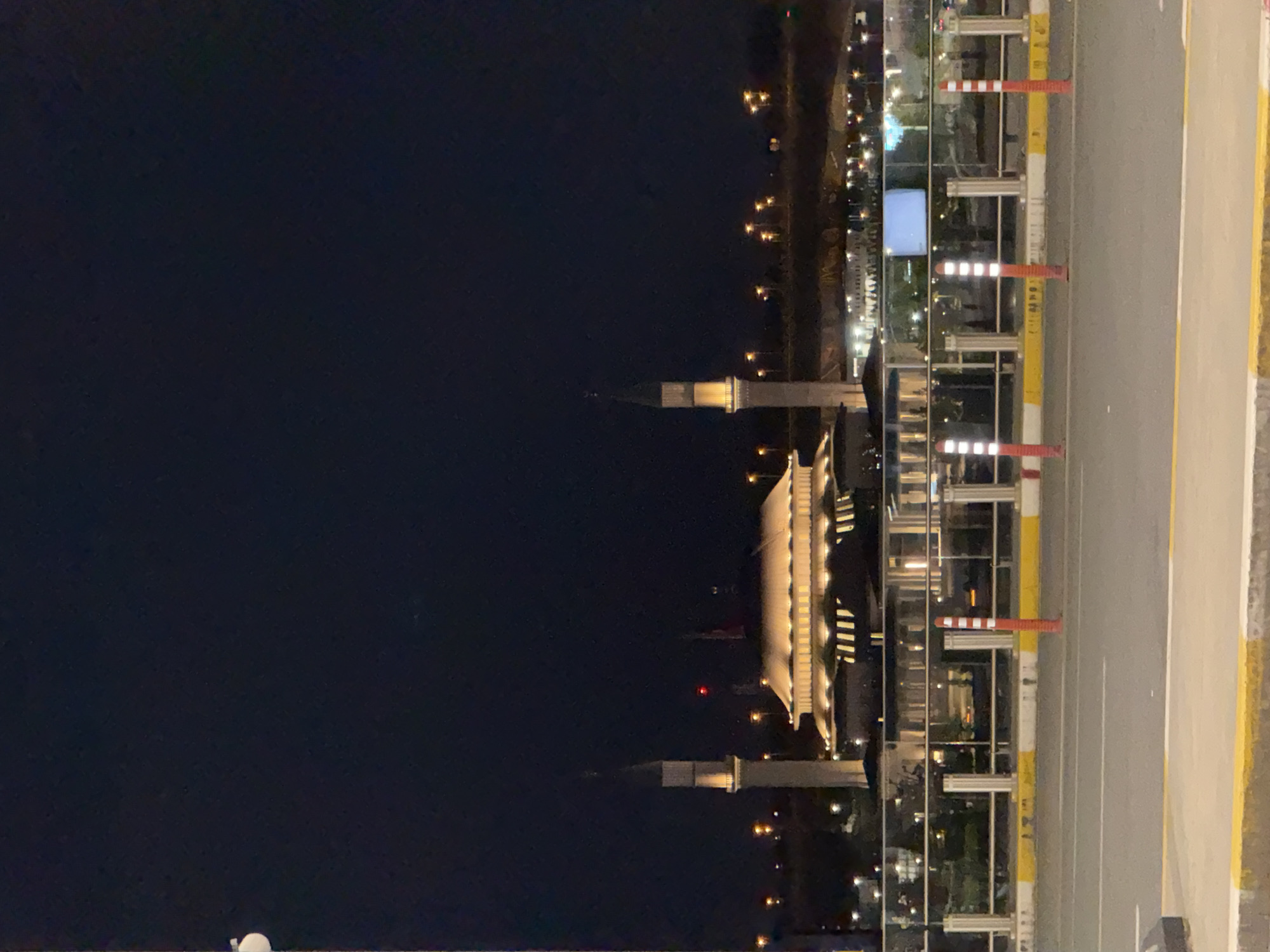 A nighttime view shows a mosque with two minarets illuminated on the bank of what appears to be a body of water, possibly the Black Sea. The scene is captured from an elevated position, looking down towards the mosque. To the right of the mosque, a road with yellow and white edge markings is visible, featuring safety barriers and traffic cones. In the distance, lights suggest urban development along the shoreline. The image location is likely in Arnavutköy, Turkey, given the architectural style of the mosque.