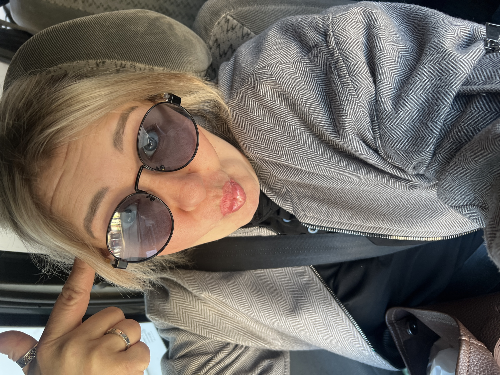 A blonde-haired individual, appearing to be female, is captured in a tilted selfie taken inside a vehicle, likely in Chaikovsky, Russia. The person is wearing round-framed sunglasses with dark lenses and a black frame, partially revealing their blue eyes. They wear a grey jacket or coat featuring a distinctive herringbone pattern, over a black shirt or top. A black seatbelt is visible across their chest, indicating they are seated in a car.
Their left hand is raised, with a finger touching their temple, and two rings are visible on their fingers: one with a dark, intricate pattern and another with small, raised dots. The person's lips are pursed in a slight pout, and their gaze is directed towards the camera, conveying a playful yet possibly contemplative expression.
The background shows the grey, textured upholstery of a car seat, and a brown bag or purse is partially visible on the lower right side. Natural light illuminates the scene, suggesting it is daytime. No discernible text is visible in the image.