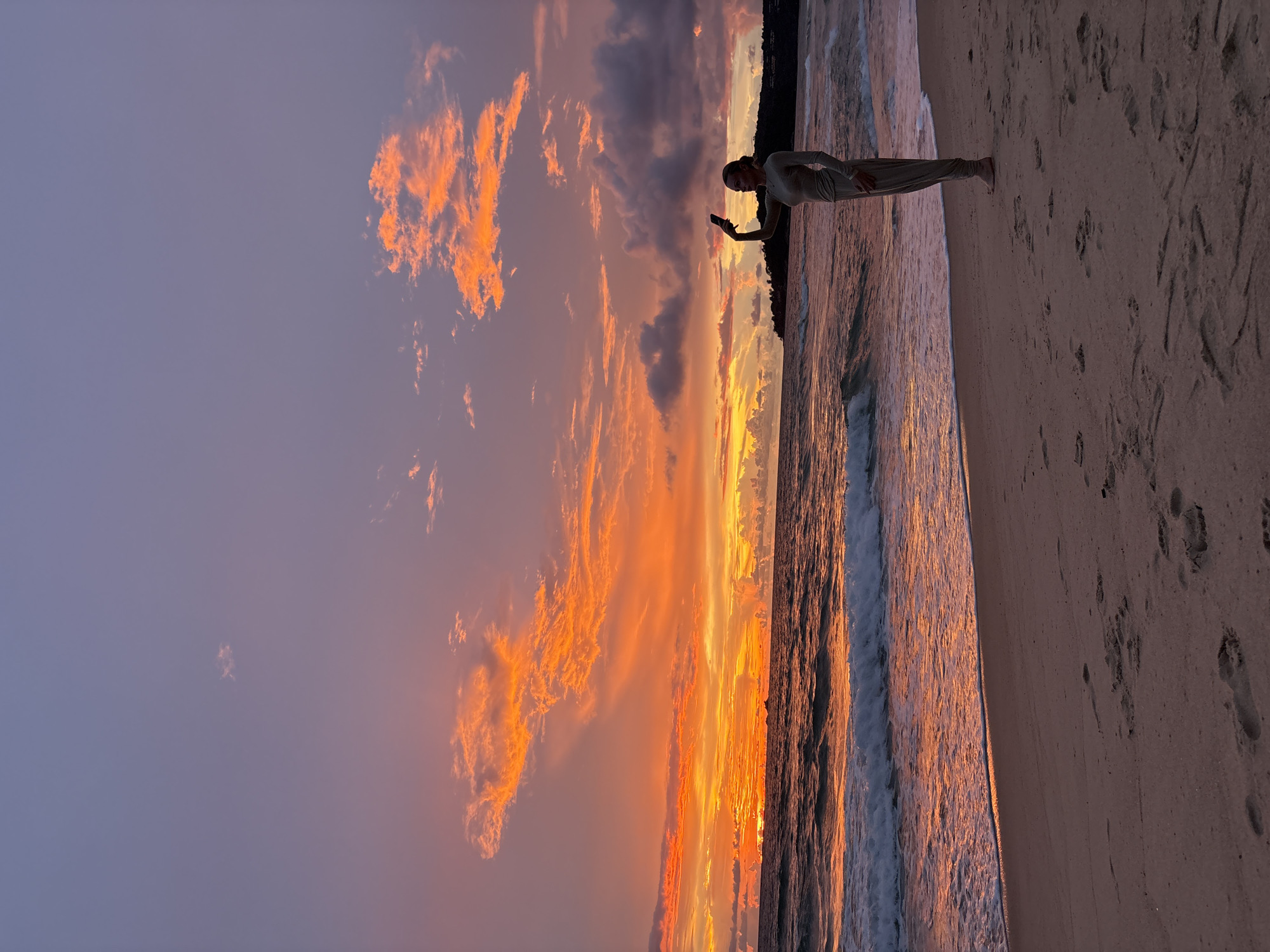 A person stands on a sandy beach at sunset, holding a phone and looking at the screen. The sky is filled with dramatic clouds painted in vibrant hues of orange, pink, and purple, indicating the sun is setting. Gentle waves lap at the shore, reflecting the warm colors of the sky. Footprints are visible in the sand, suggesting recent activity on the beach. The overall mood is serene and peaceful, with the person appearing engrossed in their phone amidst the beautiful natural spectacle. The location is Choeng Thale, Thailand.