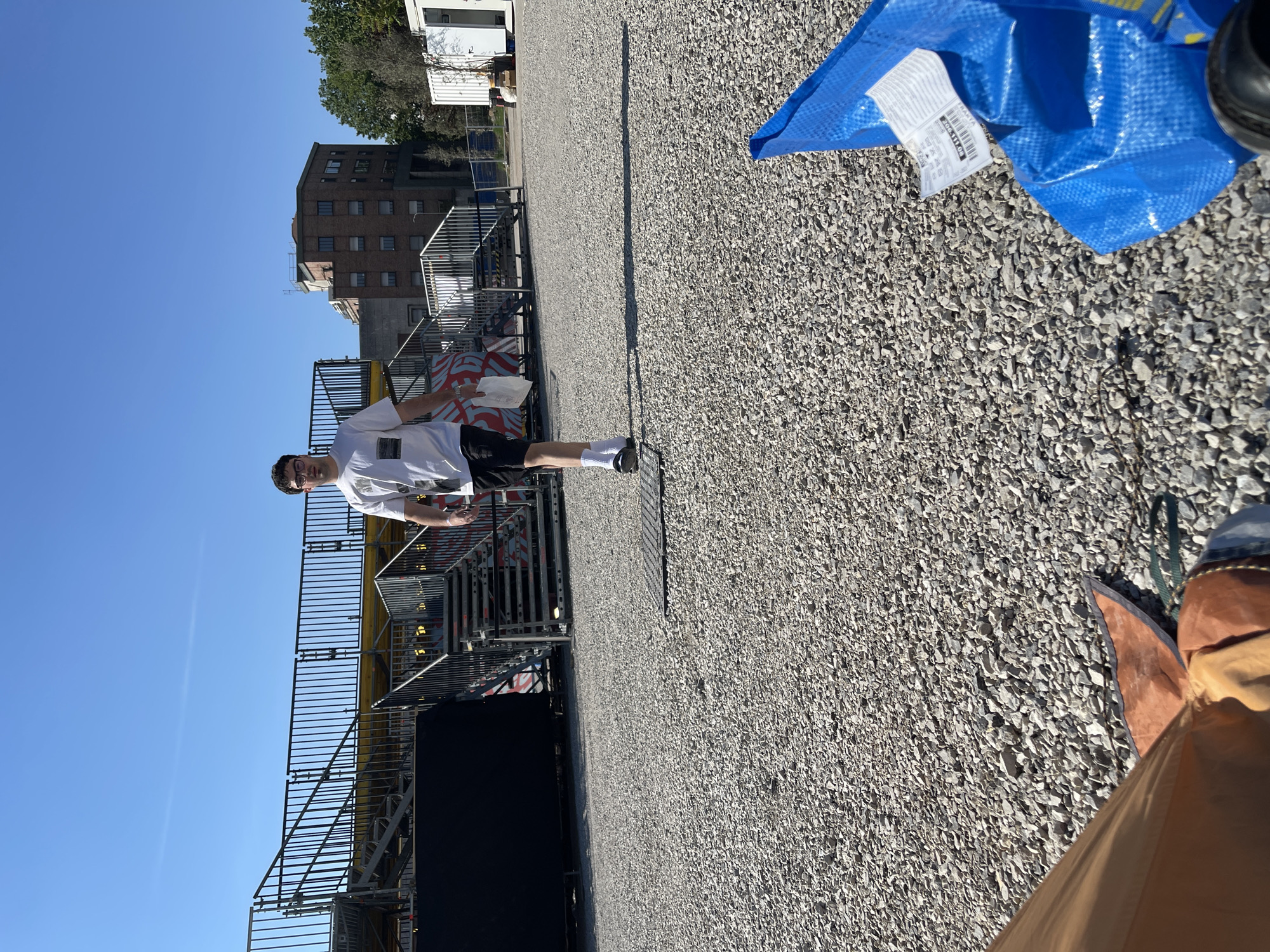 A young man with dark hair and glasses stands on an outdoor gravel surface adjacent to a metal scaffold structure. He wears a white t-shirt, dark shorts, and white socks. The sky is clear blue. A blue IKEA bag and other objects are near the edge of the frame on the right. In the background, there is a multi-story brick building and white trailers. Based on the architectural style and other visible elements, the location is consistent with a site in Milan, Italy.