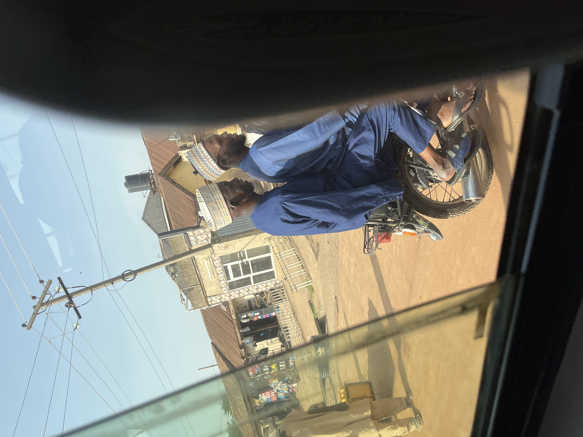 The image, originally rotated 90 degrees counter-clockwise, depicts an outdoor street scene in Ado, Nigeria, captured through a reflective vehicle window. This perspective results in a layered view, with internal reflections superimposed onto the external scene.
In the foreground, a man is seated on a black motorcycle, likely stationary. He is dressed in a vibrant blue traditional garment, possibly a kaftan, and a distinctive white and blue patterned kufi cap. His bare feet, wearing sandals, are visible near the motorcycle's footpegs. A prominent reflection of this man appears immediately to his left, creating a semi-transparent, ghost-like duplicate figure. The motorcycle features a chrome exhaust pipe and other metal components.
The background reveals a clear, bright blue sky, indicating daytime, possibly midday given the subtle shadows on the ground. The road or pathway appears unpaved, consisting of reddish-brown dirt. Several buildings line the street; one is a two-story structure with a large window, possibly a shop displaying merchandise. Another building has a reddish-brown corrugated iron roof, and a black water tank is visible on a rooftop. A tall wooden utility pole, equipped with multiple power lines and insulators, stands against the sky.
The frame of the vehicle from which the photo was taken is visible as dark bands along what would be the top and bottom edges if the image were correctly oriented. There is no discernible text visible on any of the objects or buildings. The scene conveys a typical daily moment in a local Nigerian street.
