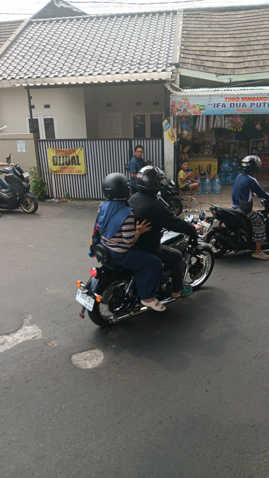 In Depok, Indonesia, on a city street, two individuals are riding a black motorcycle. The rider in the front is wearing a black helmet and a black jacket. The passenger behind them is wearing a blue hijab and a striped long-sleeved shirt, and is holding onto the rider. The motorcycle is in motion, indicated by the blur on the wheels. In the background, several other motorcycles are parked or in motion, and a small storefront is visible with the signage "TOKO SEMBAKO IFA DUA PUTRI MADURA" and "RUMAH DIJUAL" (house for sale) on a yellow banner. There are also several people standing and sitting in front of the storefront. The road surface shows signs of wear and tear with potholes. The environment suggests a typical urban scene in Indonesia with buildings, shops, and street activity.
