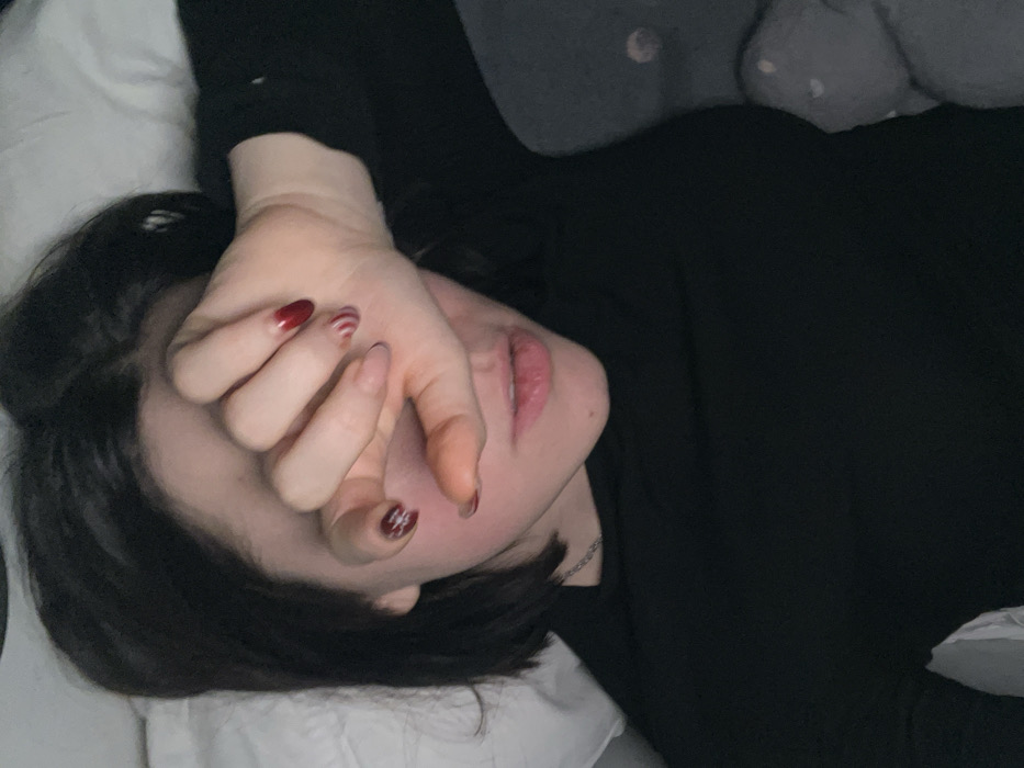 A person lies in bed wearing a black top. They have short dark hair and their face is partially obscured by their hand. The hand has manicured nails, some with red polish and some with a white swirl design. The person's lips are visible and slightly parted. Part of a white pillow is beneath their head, and a grey patterned pillow is visible in the upper portion of the image.