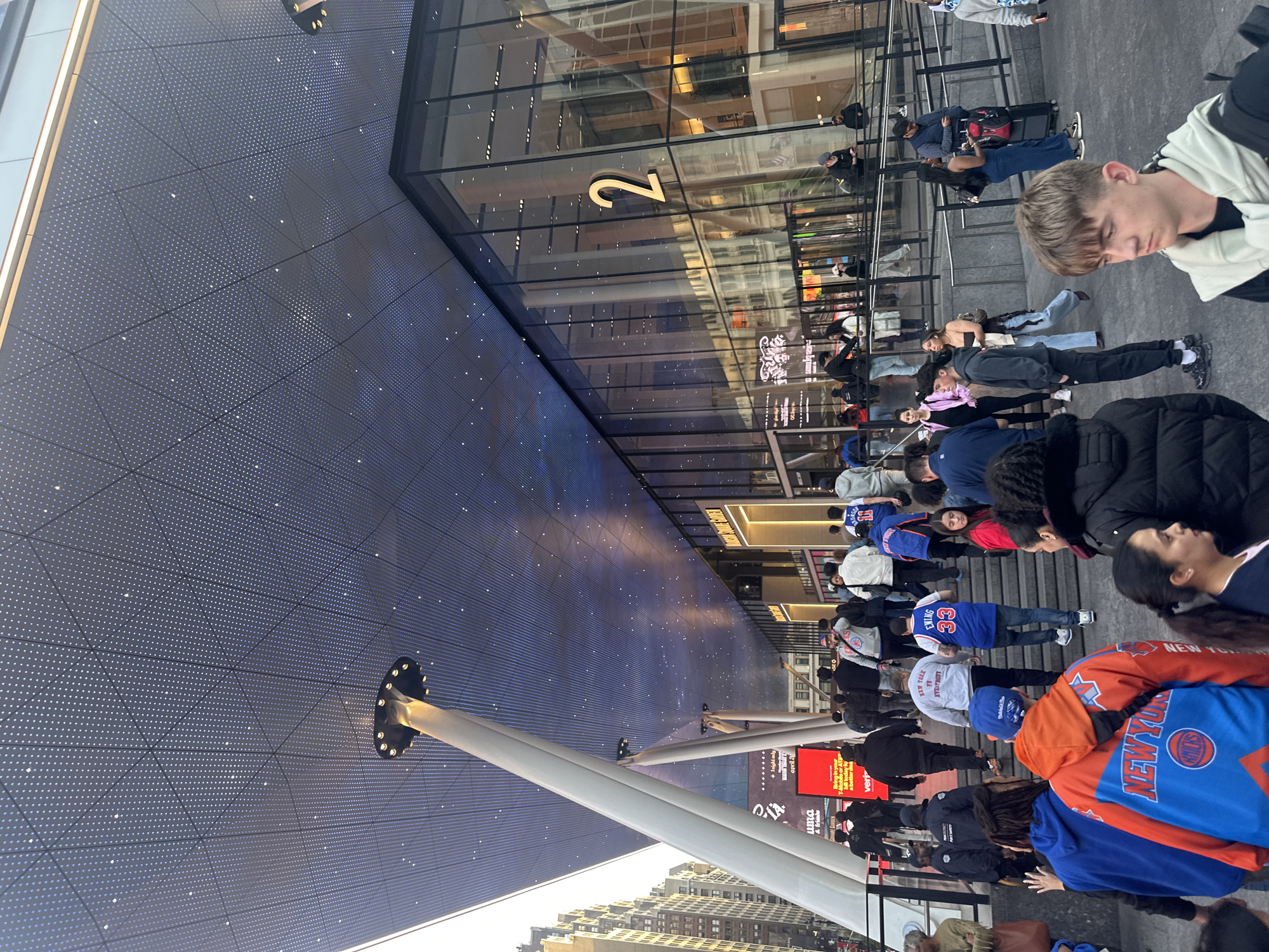 This image depicts a crowd of people gathered in front of a building in what appears to be Times Square, New York City. The building has a large, curved digital display with numerous small lights, supported by a network of white beams. The building also has a glass facade with the number "2" prominently displayed.
The crowd consists of individuals of various ages and ethnicities. Many are wearing New York Knicks apparel, including jerseys and jackets. The presence of these items and the building number suggests that they are gathered near Madison Square Garden, for some sports-related purpose, possibly for a Knicks event. People are standing on stairs, some with luggage.