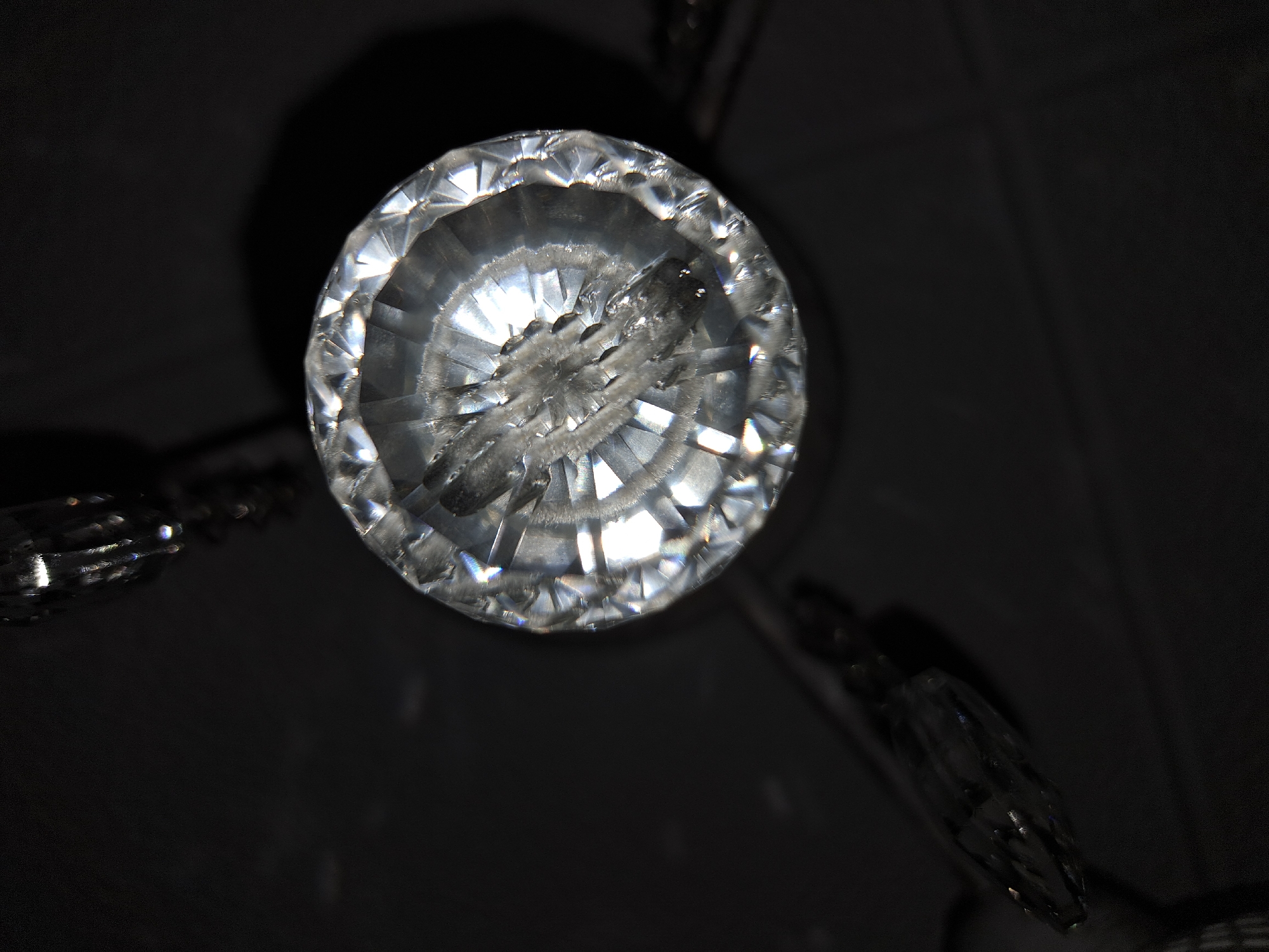 The image shows a close-up, low-angle view of a clear, round, faceted crystal chandelier component. The crystal is in focus, displaying intricate geometric cuts and internal reflections. The background is predominantly black, with dark shapes of other chandelier elements visible, suggesting a dimly lit room. There is no indication that this image was taken in Chepintsi, Bulgaria.