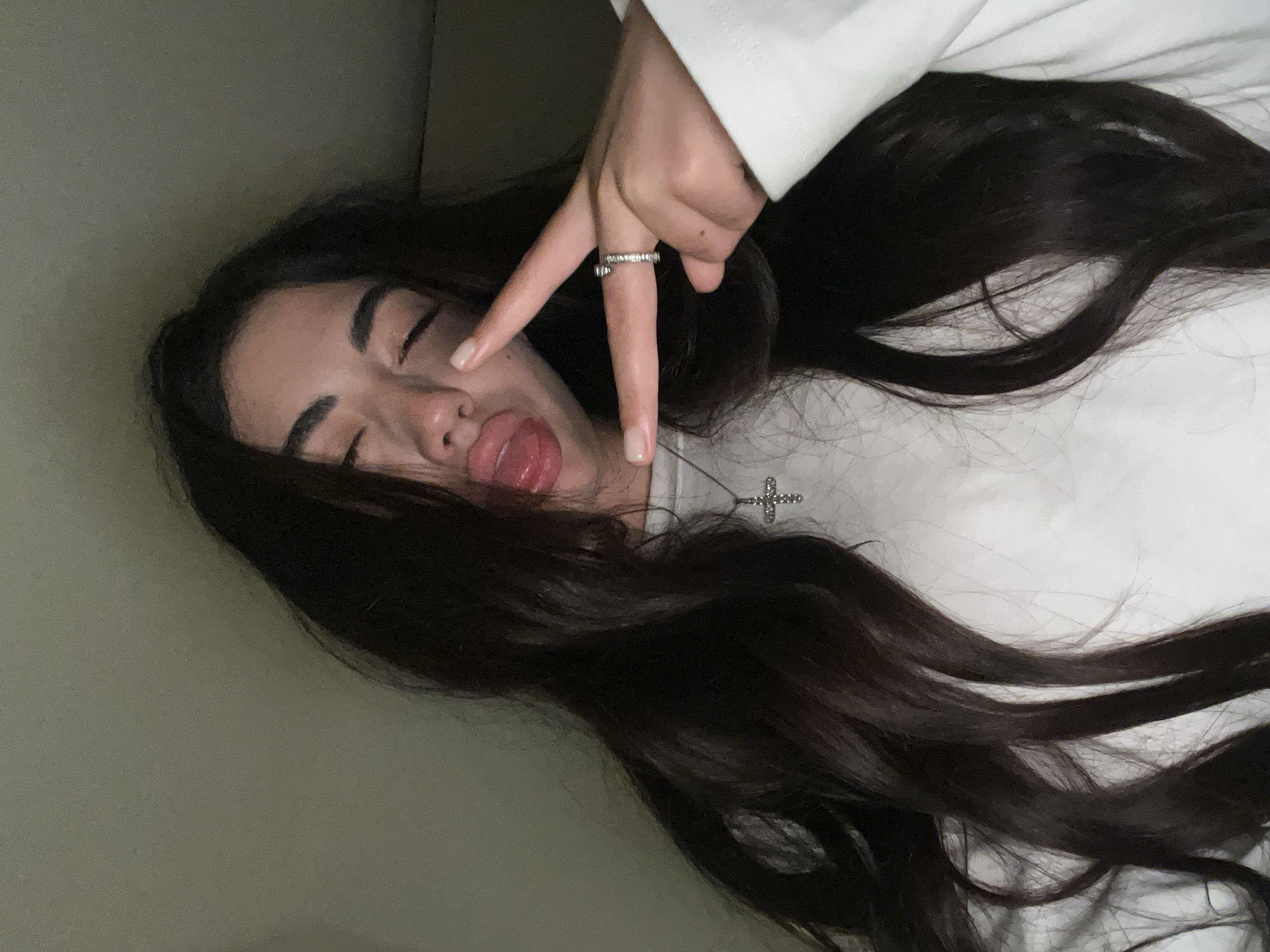 A young woman with long, dark hair is leaning forward, sticking out her tongue and making a peace sign with her right hand. She is wearing a white sweatshirt and a silver cross pendant necklace. A ring is visible on her right index finger. The background is a plain, dimly lit wall. The image appears to be a selfie taken indoors. There is no discernible information about the location in Chotěšov, Czechia.