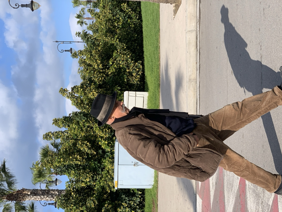 The image captures an adult male walking across a paved surface in Tangier, Morocco. The man, appearing middle-aged with a graying mustache, wears a black fedora with a beige band, a brown quilted puffer jacket, a dark blue undergarment, a dark scarf, and light brown corduroy trousers. His hands are in his jacket pockets, and he casts a distinct shadow to his left, indicating direct sunlight. Red and white markings are visible on the paved surface in the lower left. A light gray curb separates the paved area from a strip of green grass, where a light blue utility box and a white utility box are situated behind the man. The background features dense green foliage, including shrubs and several tall palm trees, beneath a clear blue sky with scattered white clouds. Multiple ornate black streetlights are visible amongst the greenery.