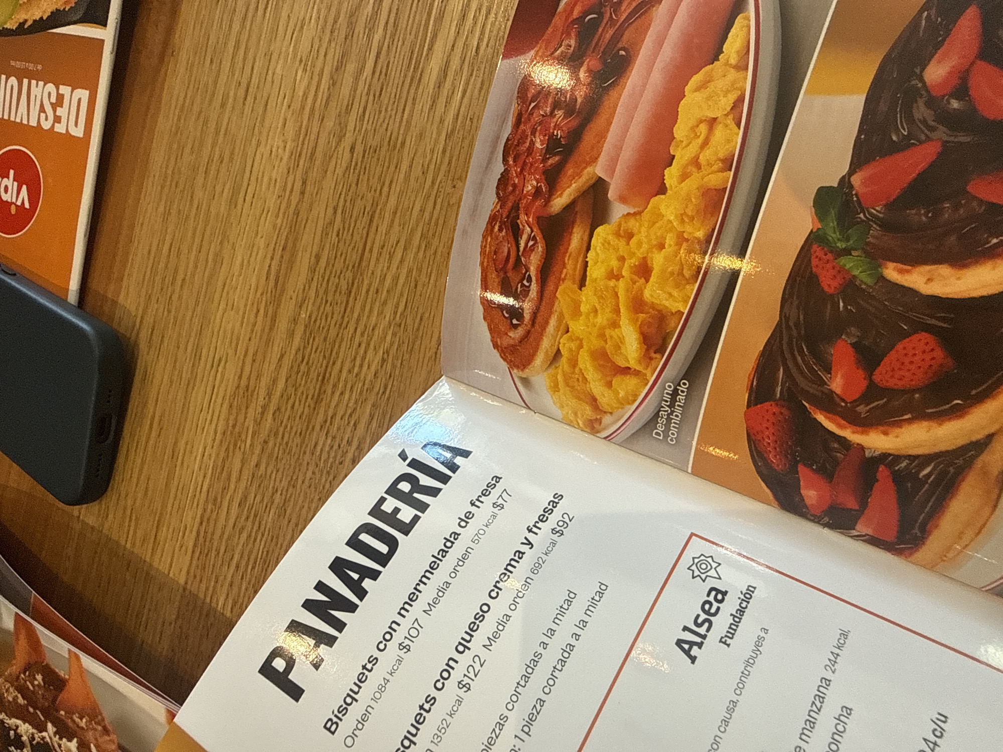 Here is a forensic description of the image:
The image shows a menu from a restaurant, showcasing the "Panadería" section, with descriptions of food items. The text is in Spanish, and the menu offers "Bísquets con mermelada de fresa" (Biscuits with strawberry jam) and "quets con queso crema y fresas" (quets with cream cheese and strawberries), including prices and calorie information. A portion of a breakfast combination meal is pictured above the menu items, featuring scrambled eggs, bacon, ham, and what appears to be chocolate-covered pancakes with strawberries. To the left of the menu is a black smartphone on a wooden surface. A portion of another menu is also visible with a "Vip" logo, partially showing "DESAYU". The Alsea Fundación logo is visible, indicating that the menu items contribute to a charitable cause.